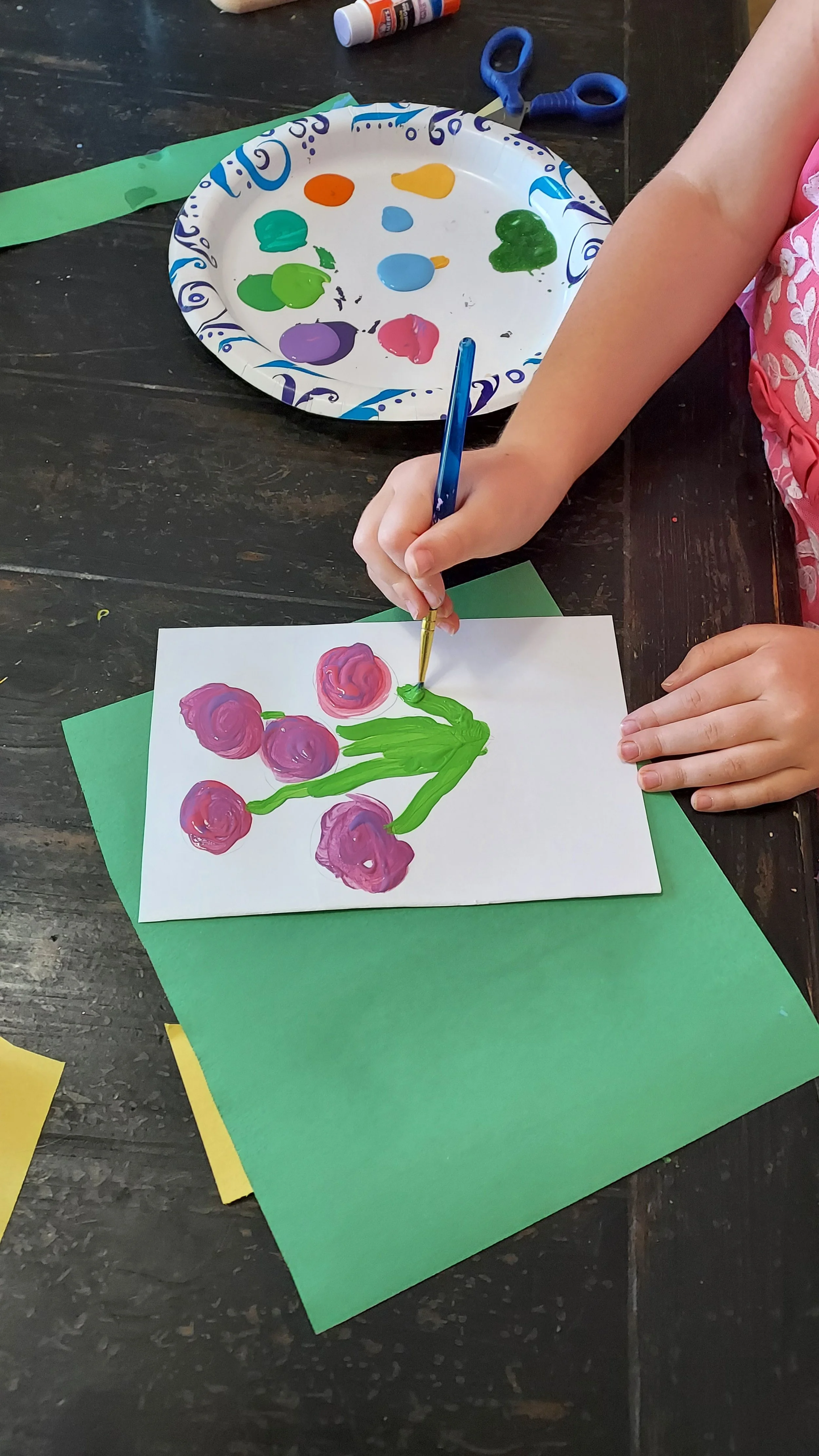 Child's hands painting a picture of a pink and purple flower with green leaves on white paper, surrounded by craft supplies like scissors, glue, and colored paper.