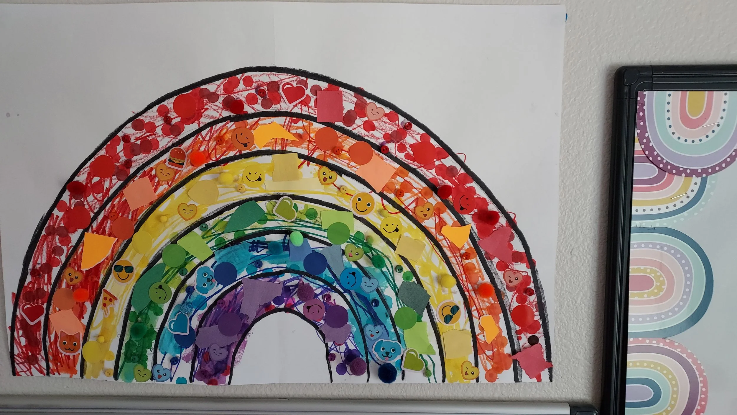 Colorful children's rainbow art with black outlines and various stickers, pom-poms, and decorations in rainbow colors, displayed on a white wall with part of a rainbow pattern on a black-framed whiteboard to the right.