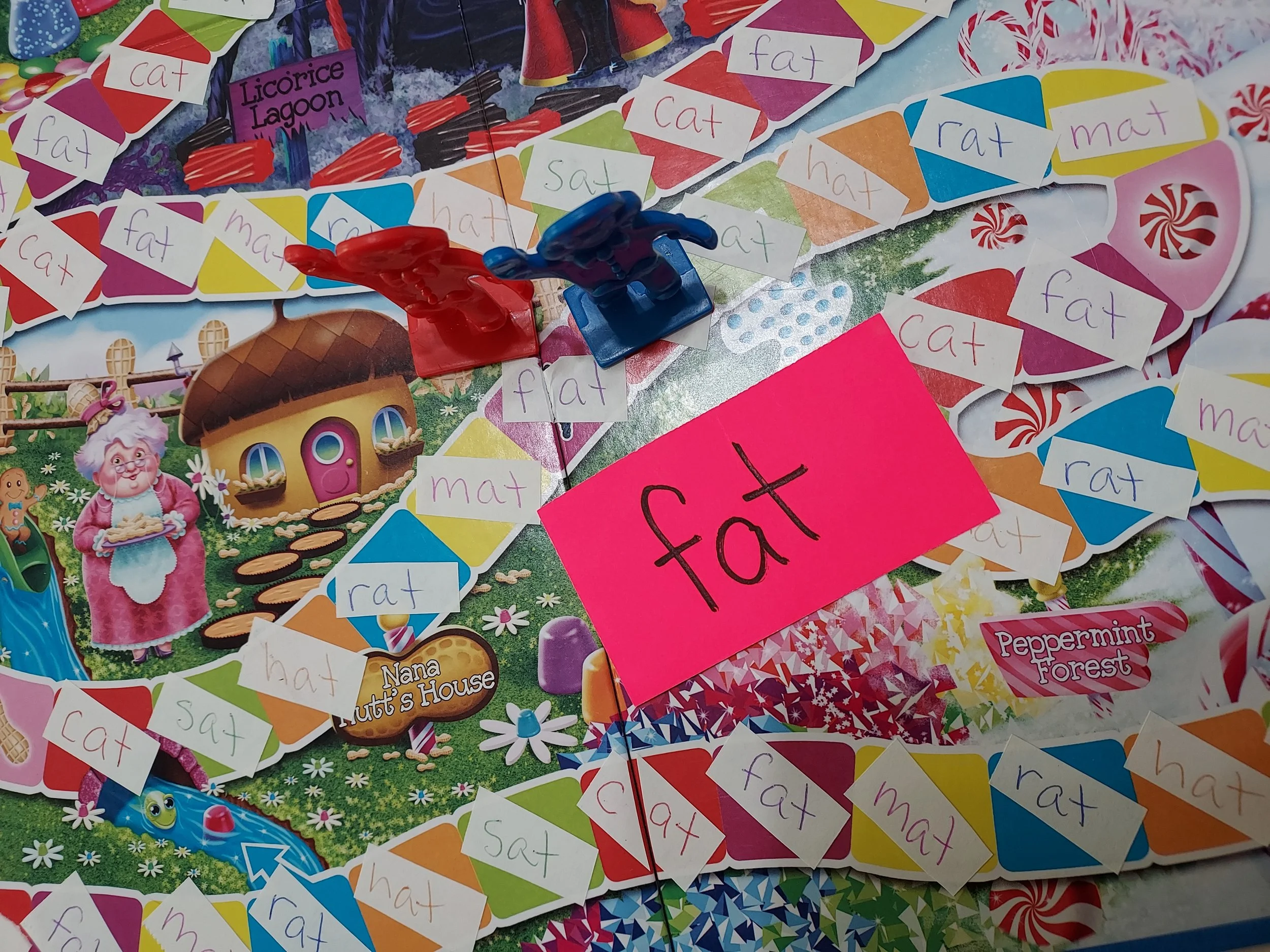 Colorful board game featuring various words like 'cat,' 'rat,' 'hat,' 'fat,' and 'mat' on small paper pieces, with two plastic player tokens, one red and one blue, and illustrations of a whimsical house, an elderly woman, and a green dragon.