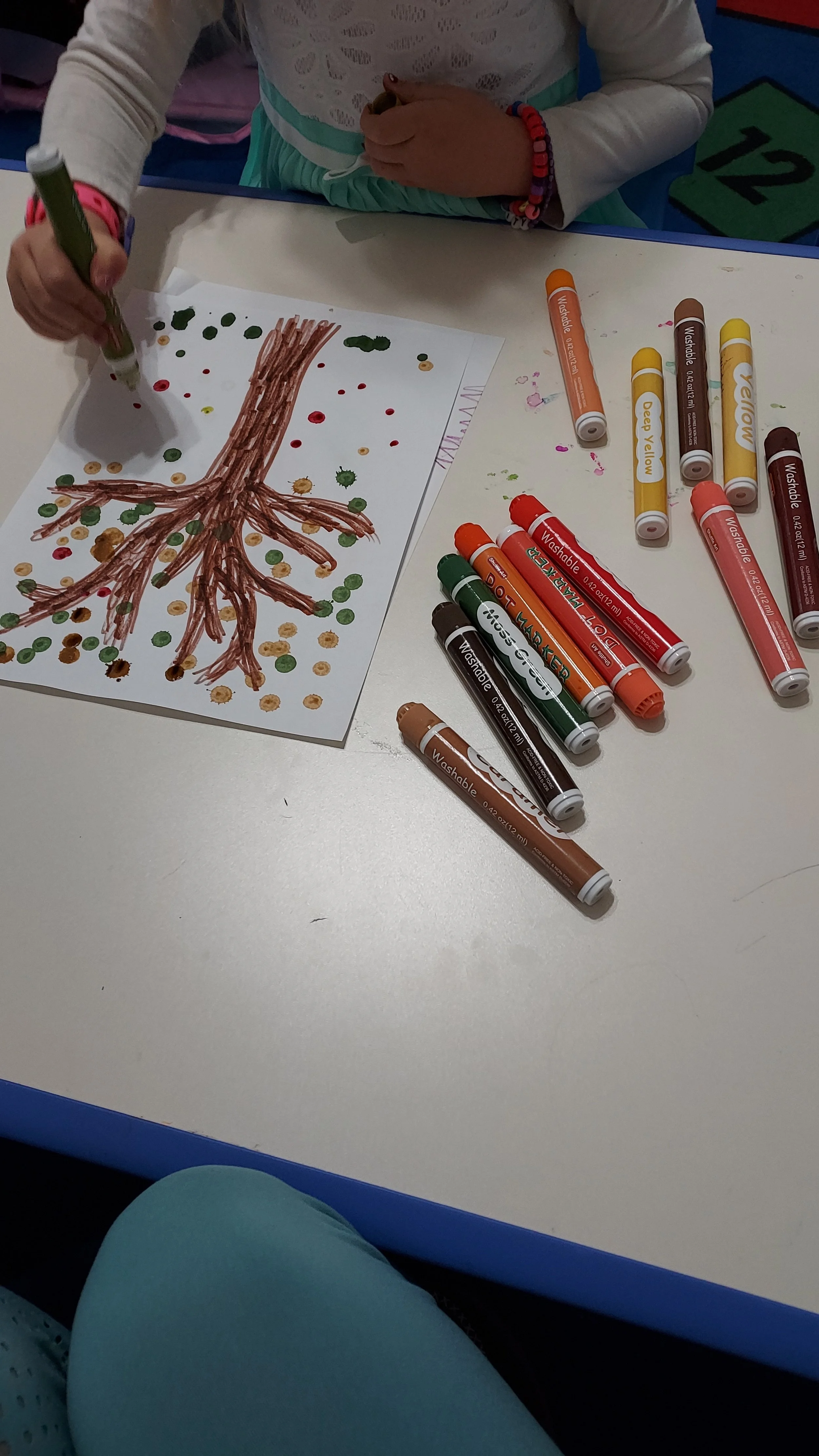 Child creating a dot art tree on paper with brown, green, red, yellow, orange, pink, beige, and brown markers, surrounded by various colored markers on a white table.