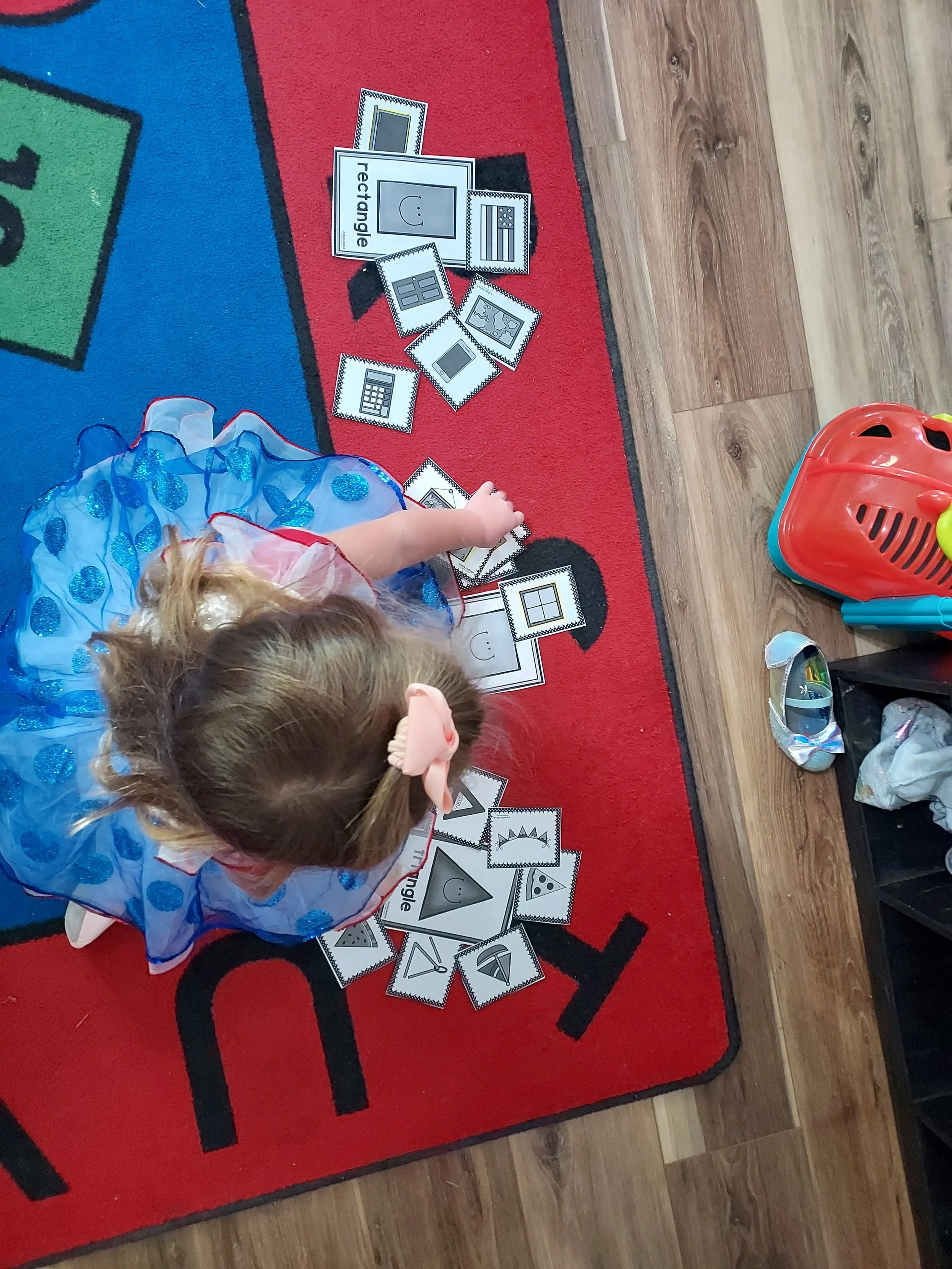 A young girl in a blue dress with a pink bow on her hair is sitting on a red carpet, playing with flashcards that have various shapes and faces, spread out on the carpet.