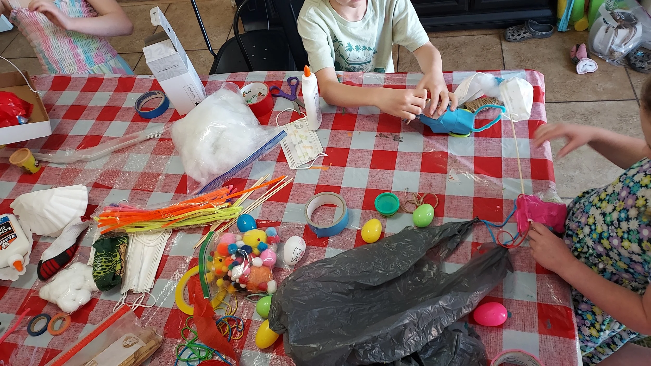 Children engaging in arts and crafts activities at a table covered with glue, tape, scissors, colorful plastic eggs, pom-poms, and craft supplies.