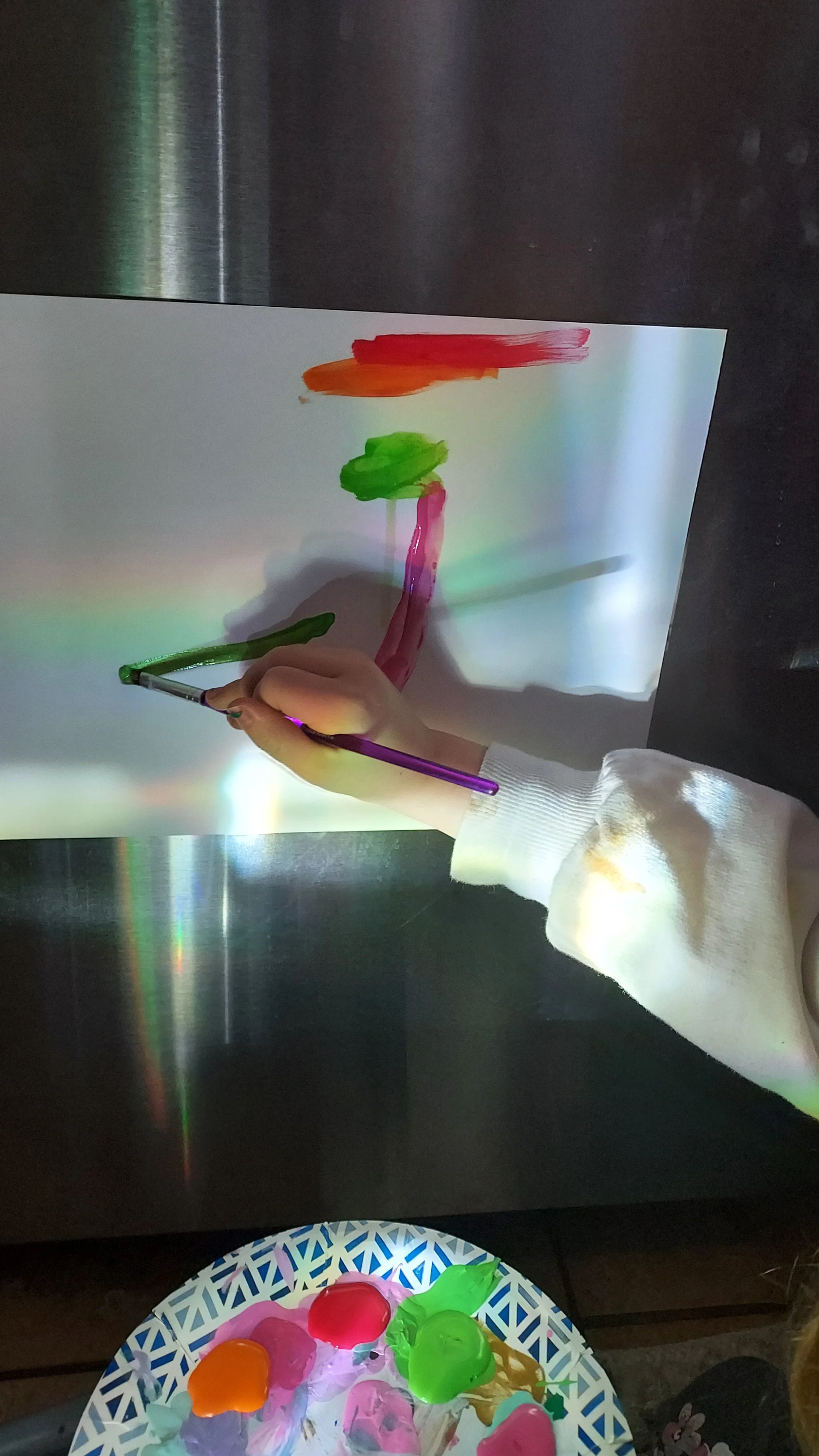 Person painting on a white paper with colorful paint, using a purple brush. The paper is tilted, and the person's hand is visible, wearing a white sleeve. A plate with various colorful paint blobs is on the table below.