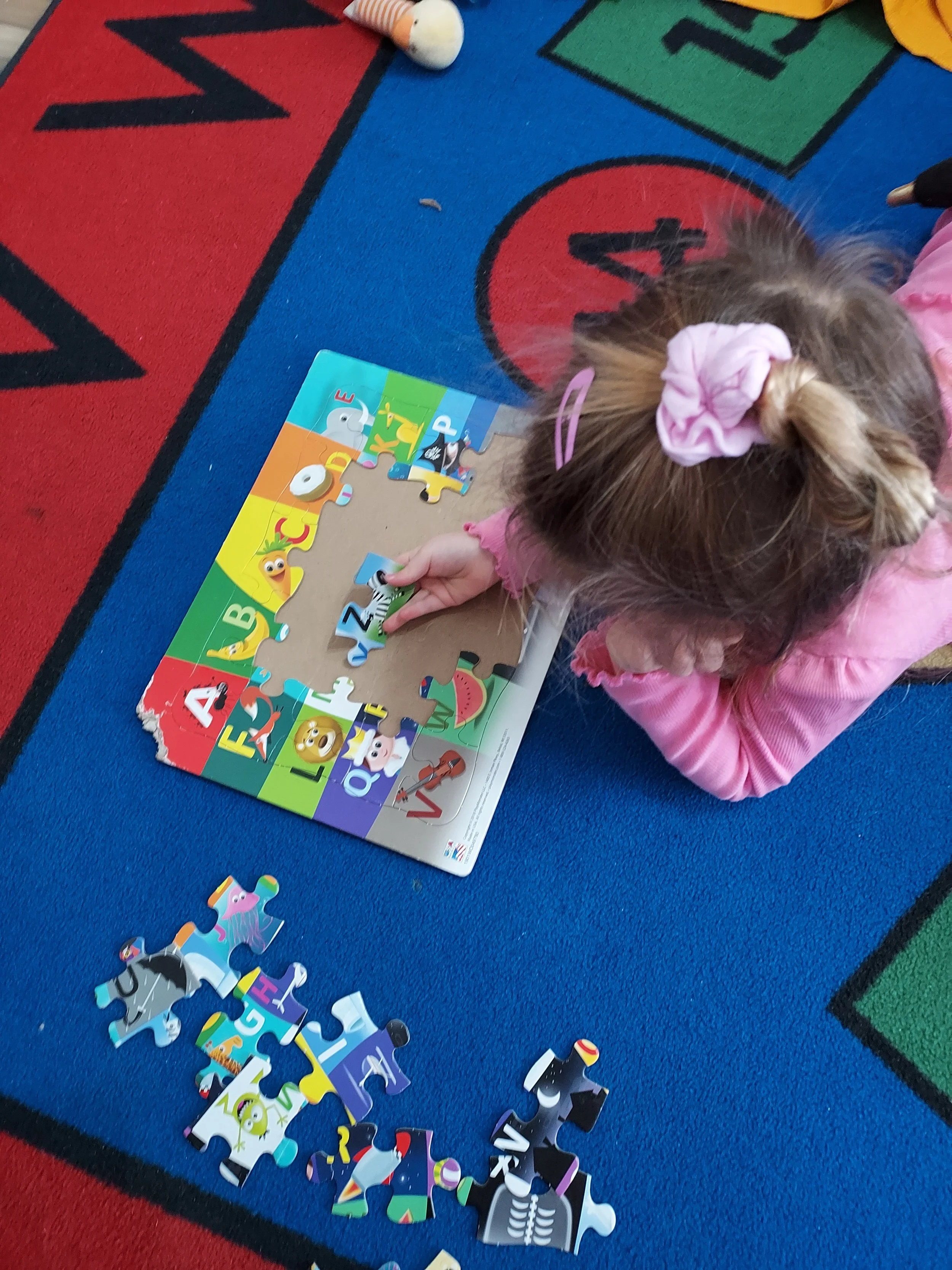 A young girl with a pink scrunchie in her hair is sitting on a colorful alphabet-themed rug, assembling a children's puzzle featuring animals, letters, and numbers.