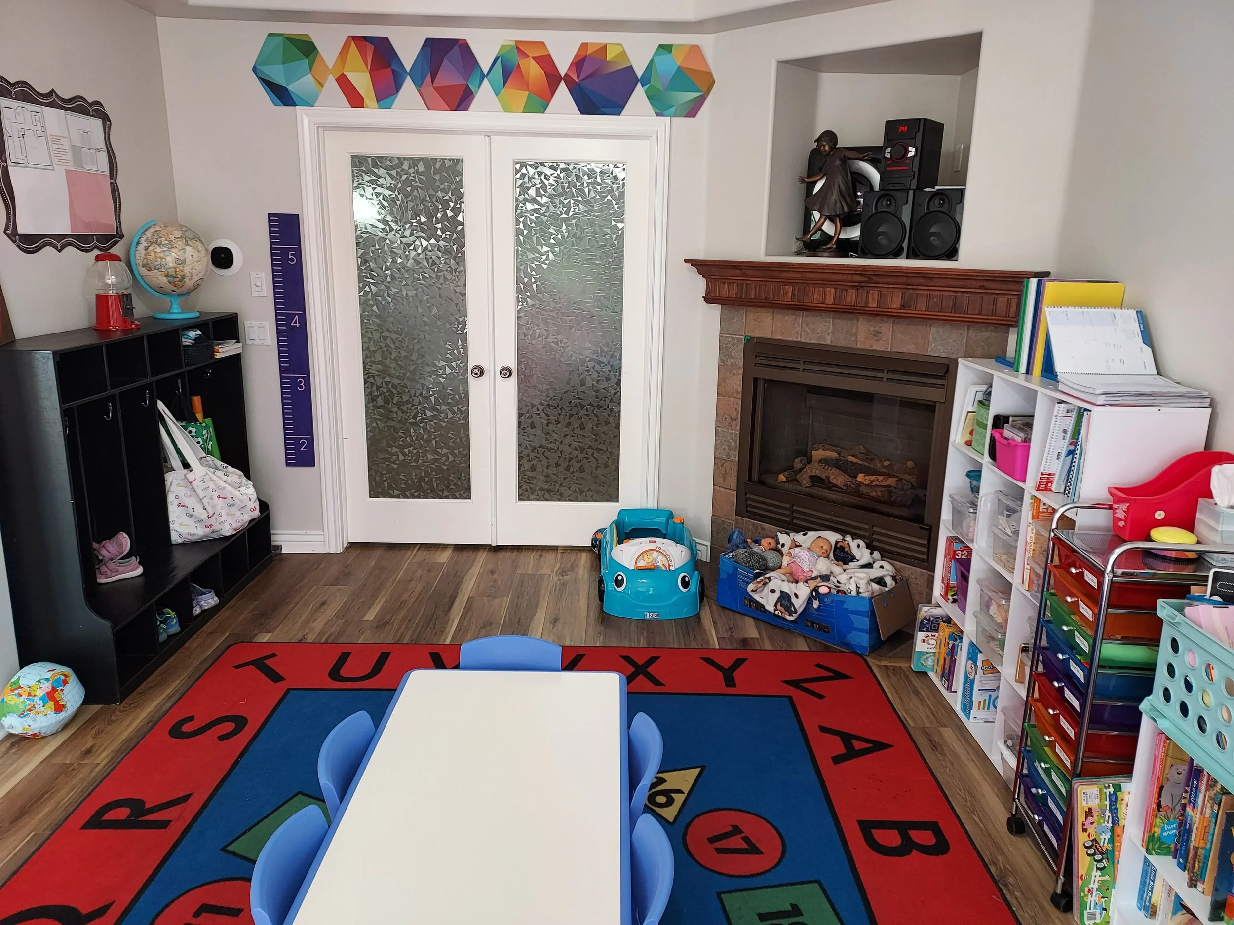 A children's playroom with a colorful alphabet rug, a white table with blue chairs, a bookshelf filled with toys and books, a fireplace with a box of plush toys, and a small toy car on the floor. A door with frosted glass leads outside, and a height measurement chart is on the wall. There are shelves with storage bins, a globe, a gumball machine, and a wall decoration made of colorful geometric shapes.