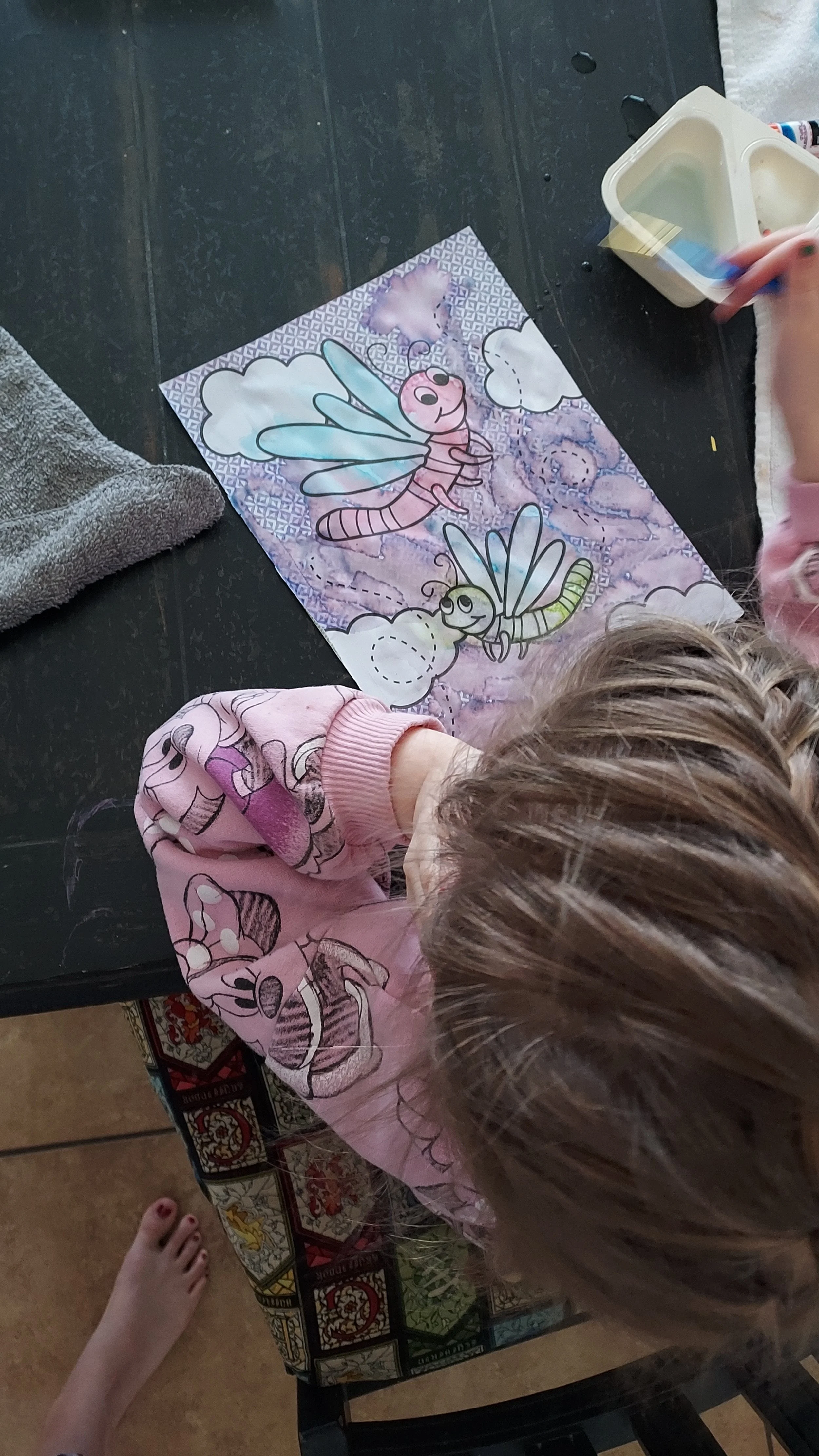 Child coloring a butterfly and bee with markers on a worksheet featuring clouds and a purple background.