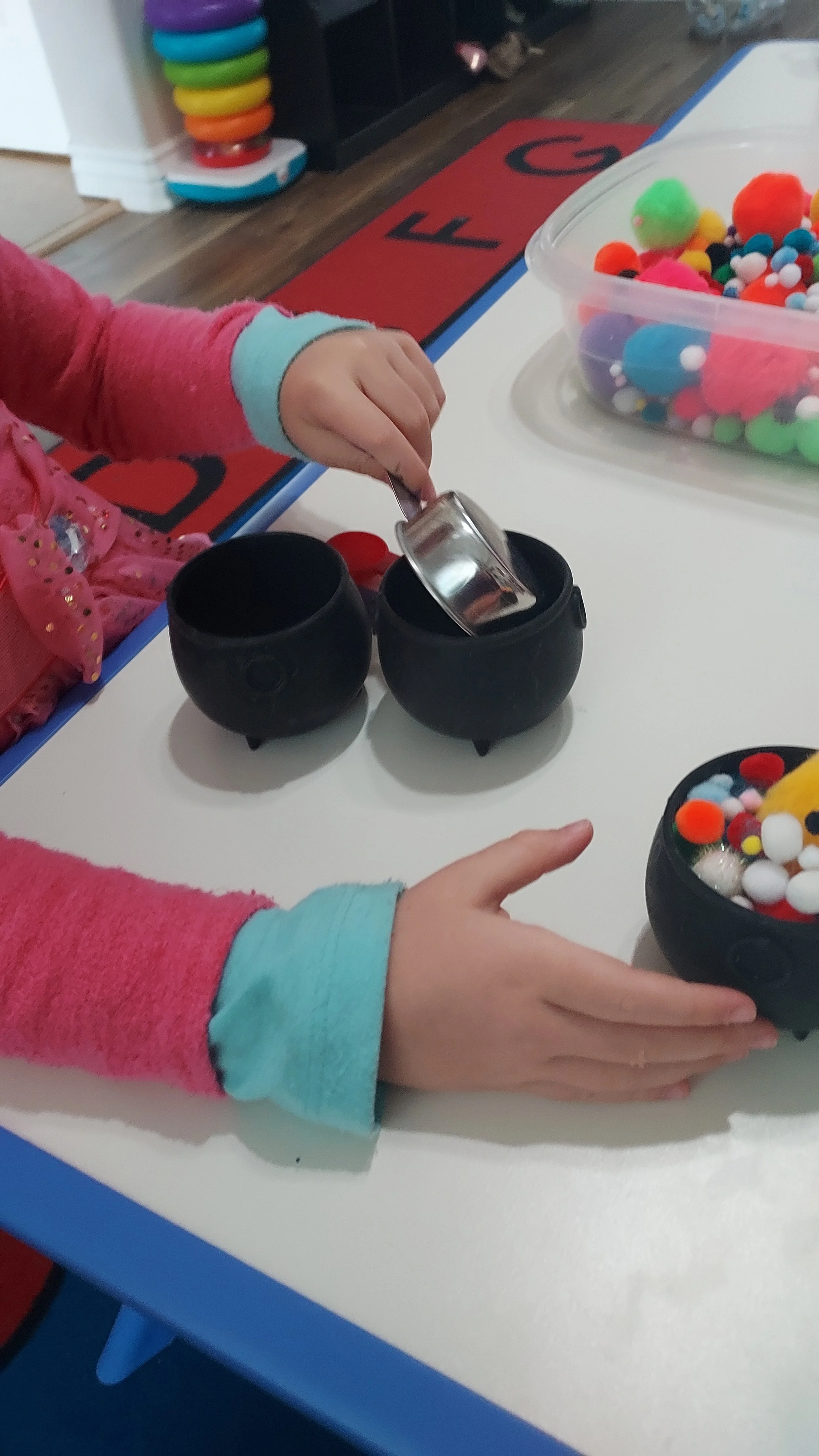 Child's hands pouring contents from a metal cup into a black textured container, with a clear container of colorful pom-poms nearby on a white table.