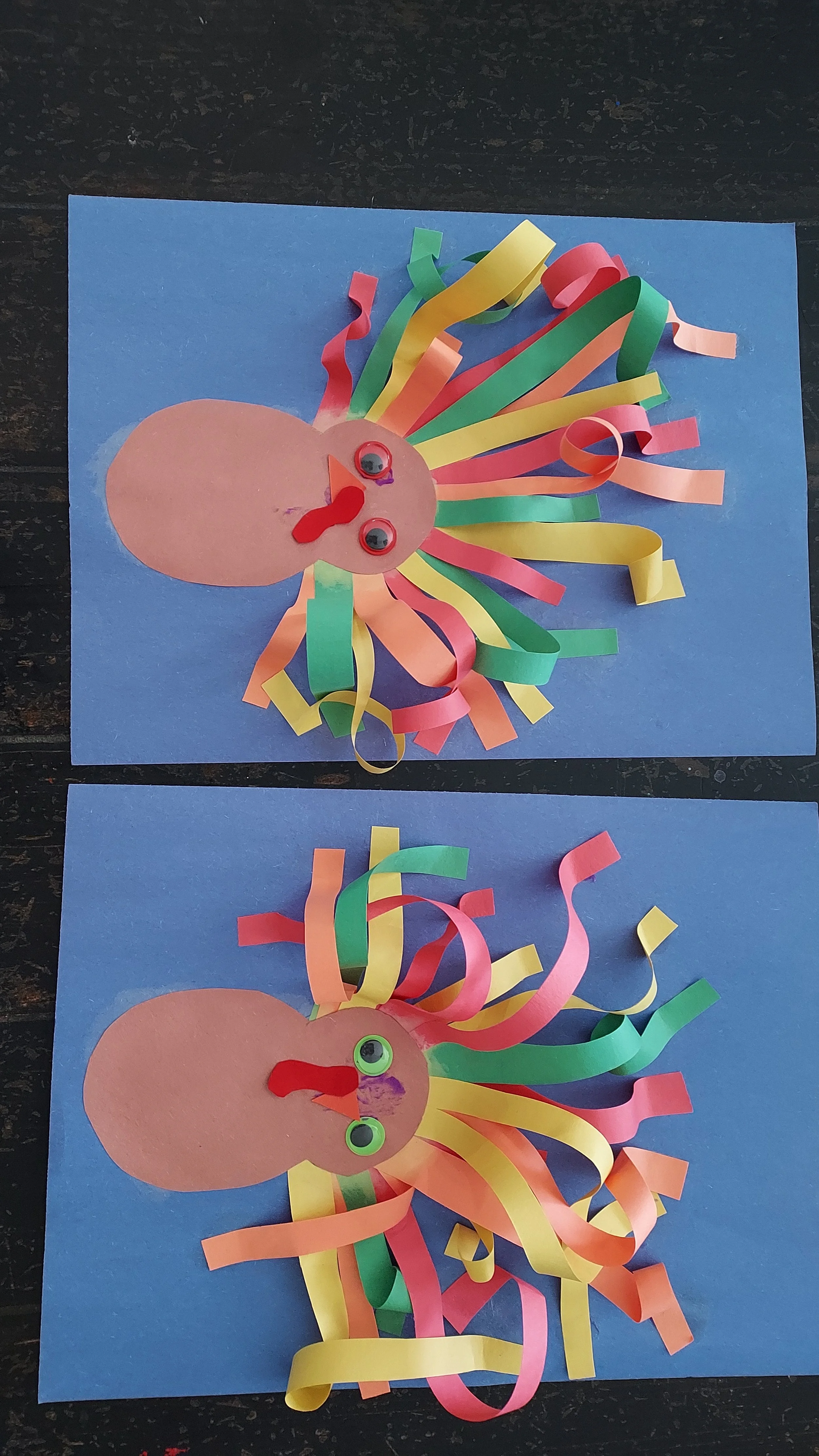 Colorful paper crafts resembling turkeys with paper plate heads, googly eyes, and curled rainbow-colored paper strips as feathers on blue backgrounds.