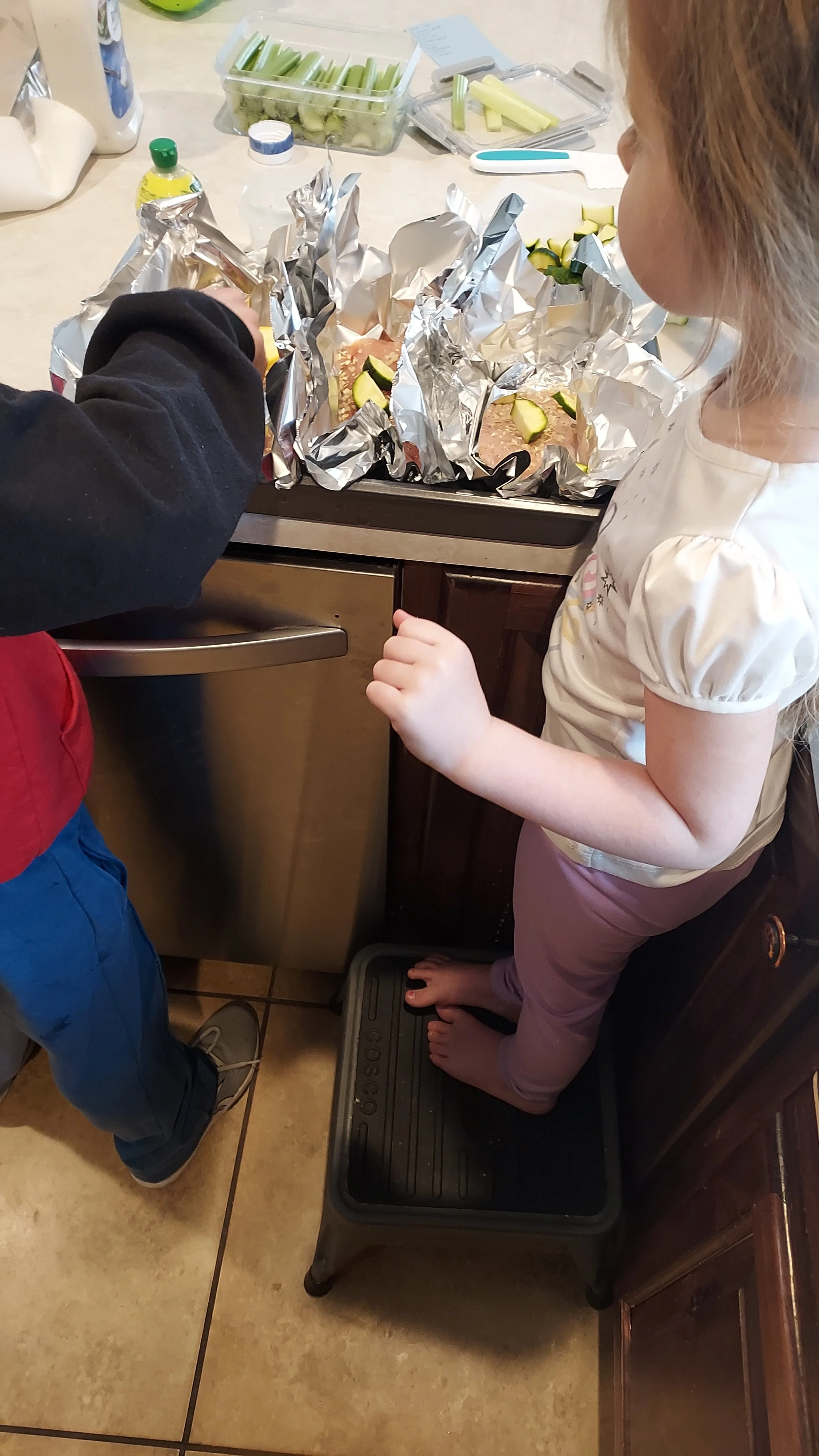 Two children prepare foil packets with zucchini and chicken in a kitchen.