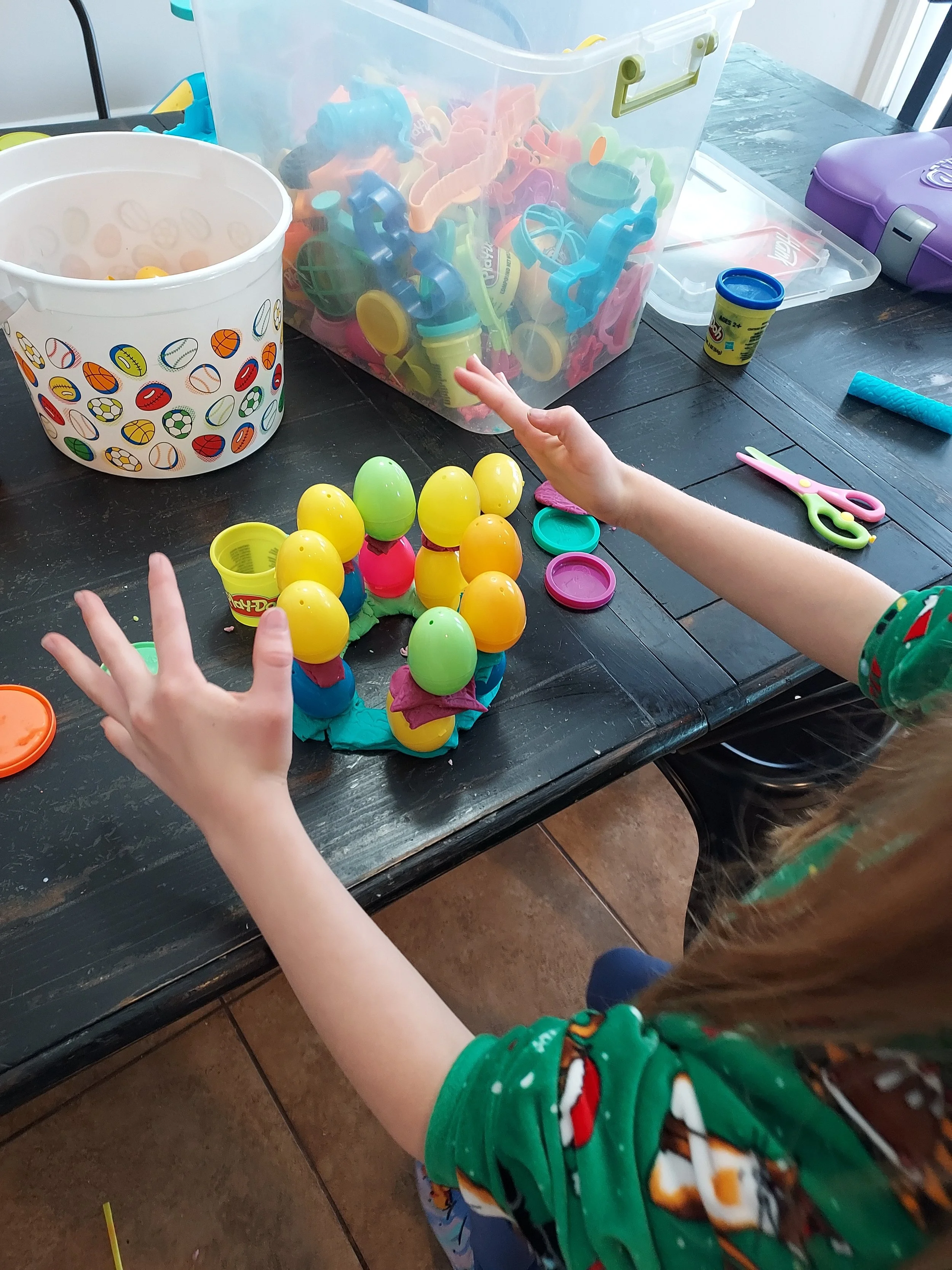 Child playing with colorful plastic eggs and toy eggs on a black table, surrounded by toys, scissors, and containers.