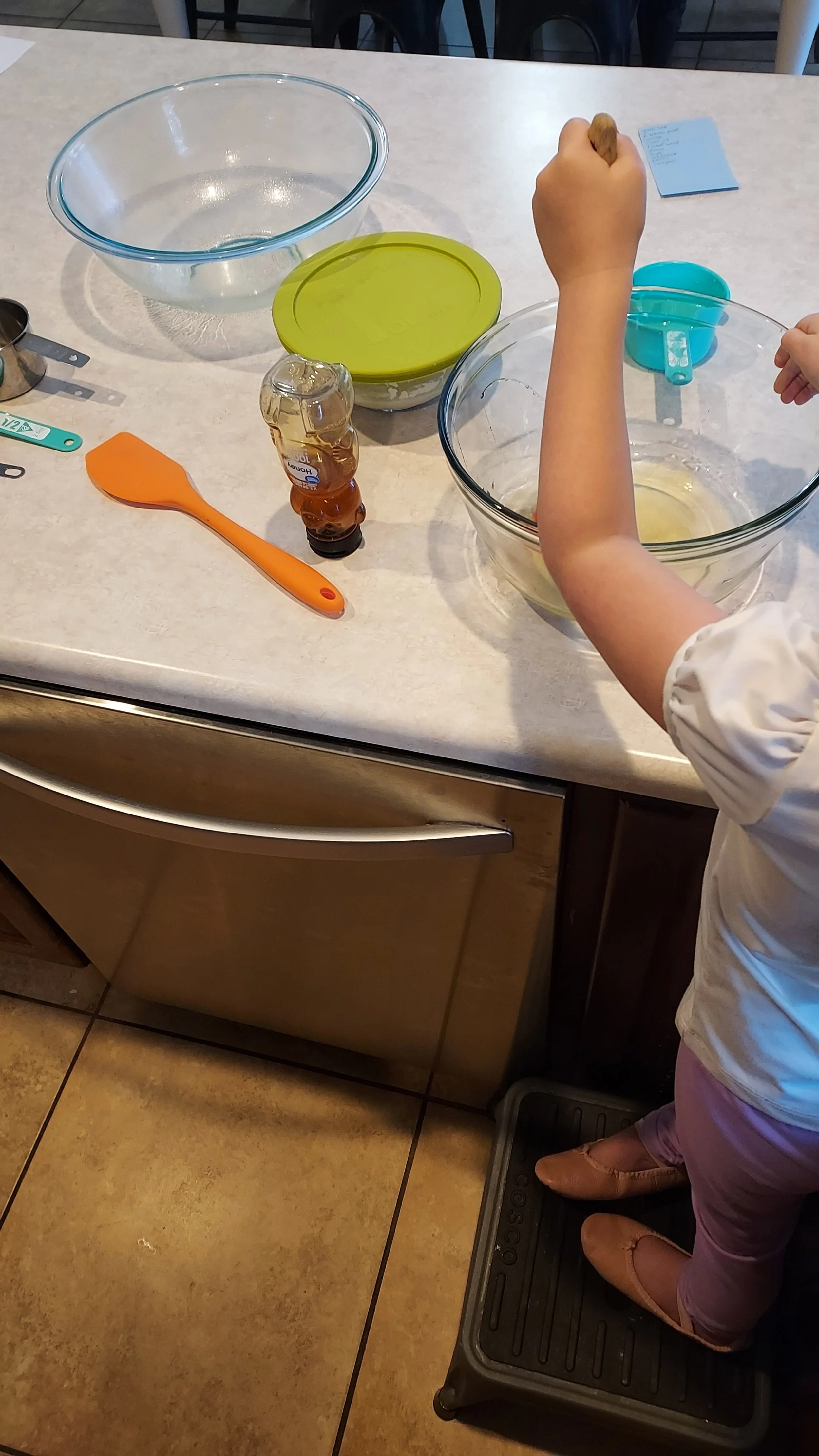 Child baking in the kitchen with mixing bowls, measuring spoons, and a small bottle of vanilla extract on the countertop.