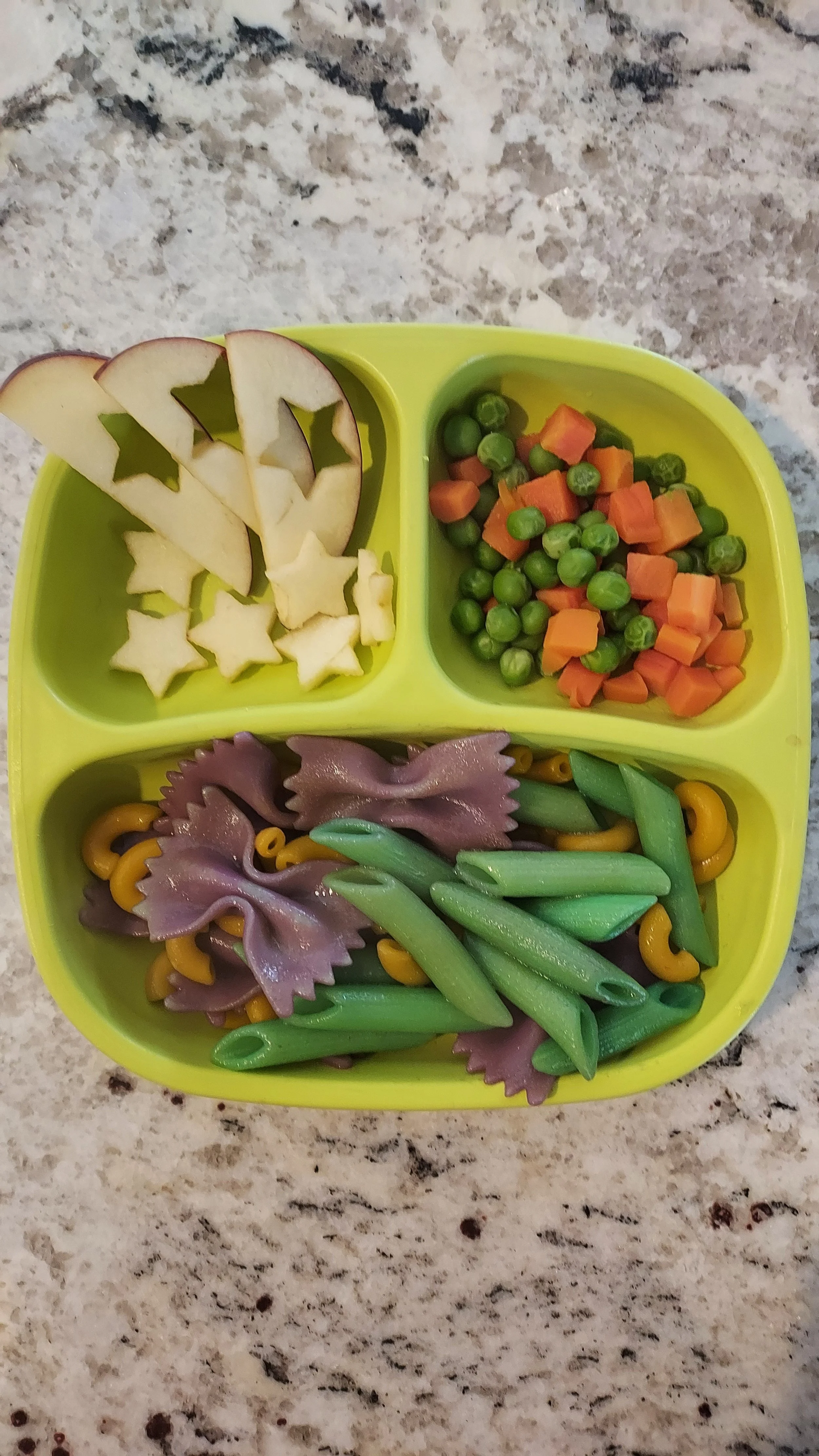 A yellow divided plate with sliced apple and star-shaped cheese in one section, green peas and diced carrots in another, and rotini pasta with purple bowtie pasta and green beans in the last section, placed on a granite countertop.