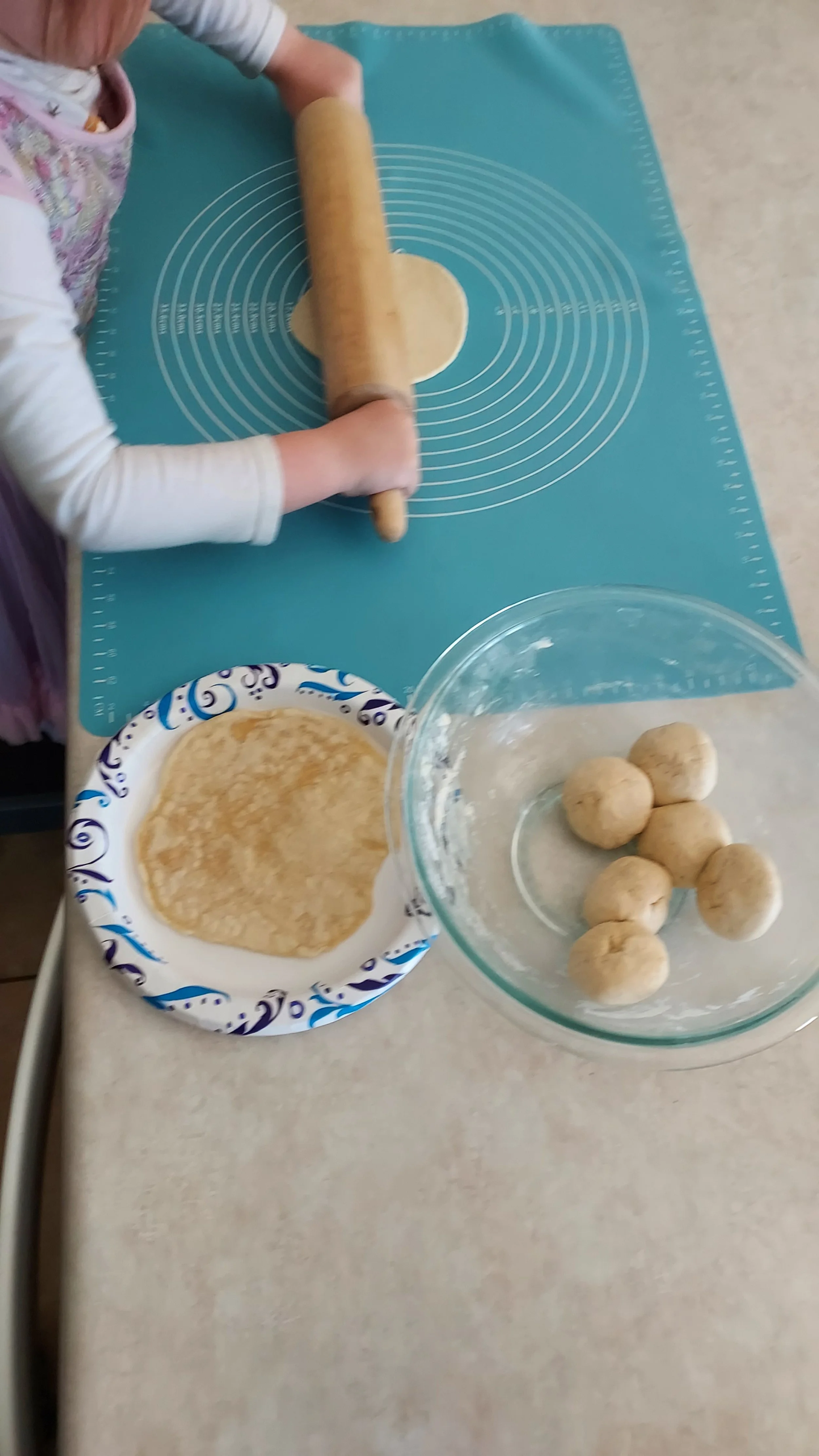 Child rolling out dough on a teal silicone baking mat, with a paper plate holding more dough balls and a glass bowl of round dough balls nearby.