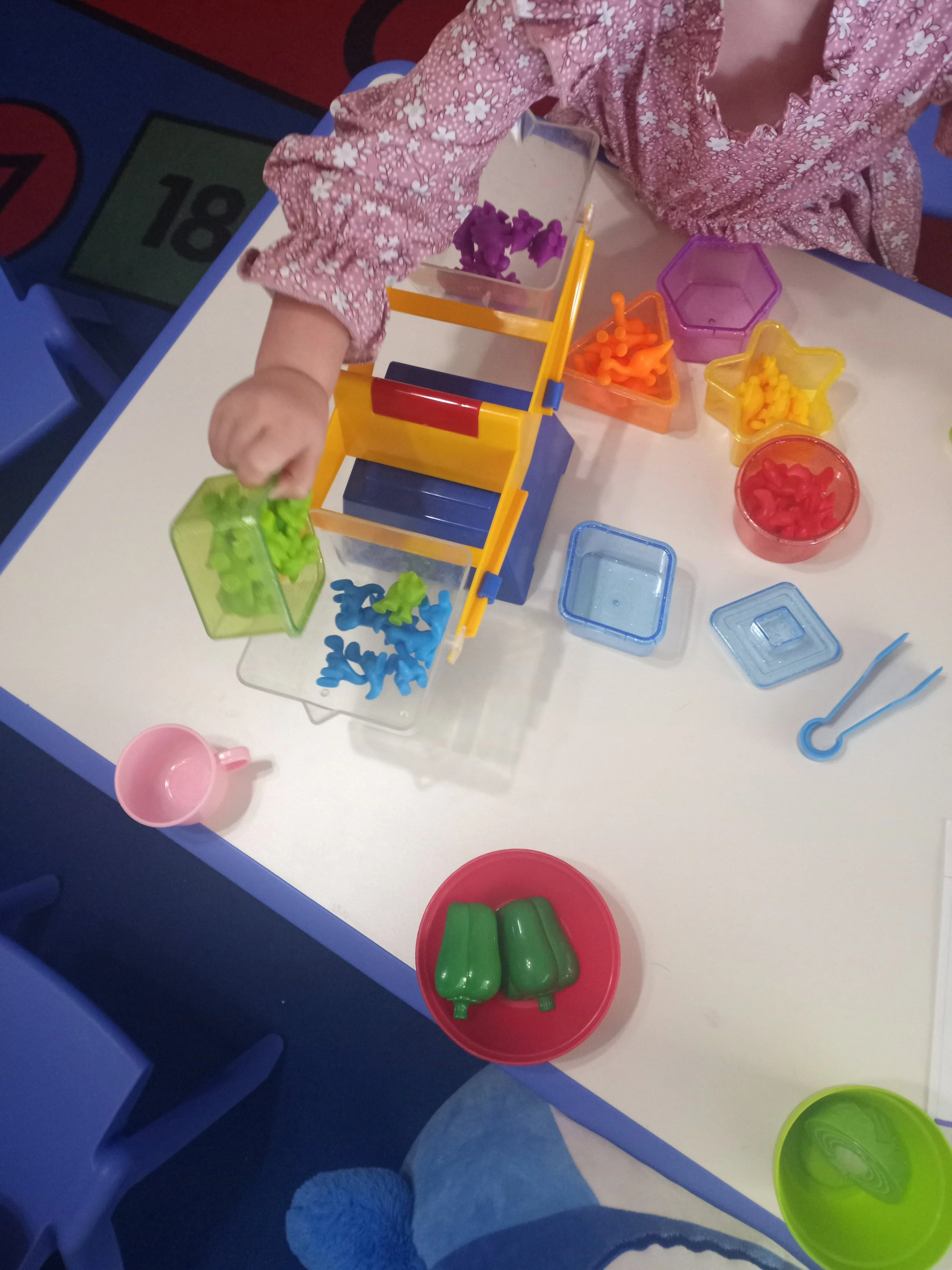 Child playing with colorful plastic toy construction set, with various parts like green, blue, yellow, orange, purple, and red pieces on a white table.