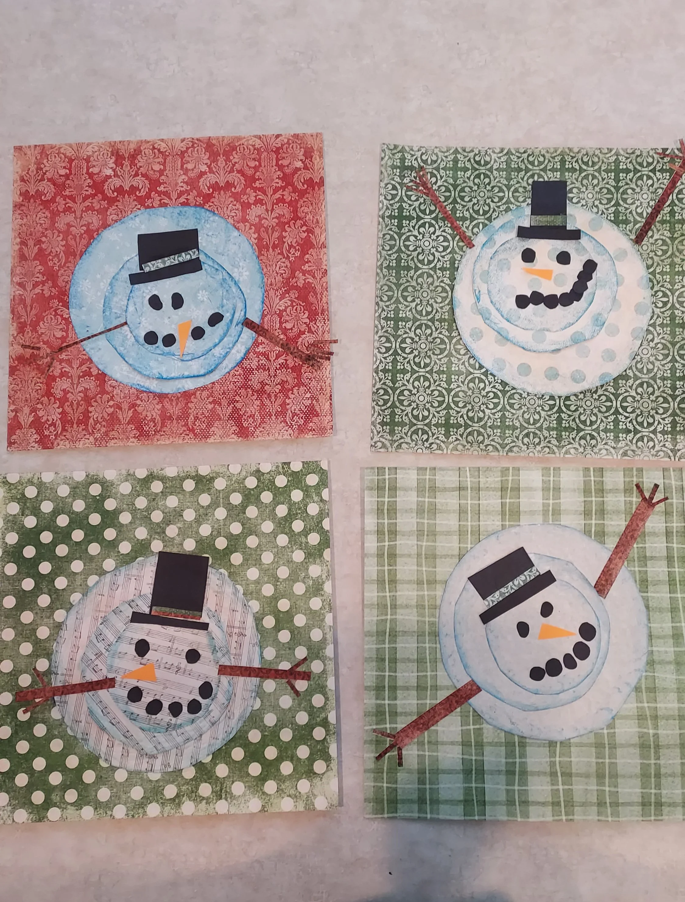 Four handmade snowman crafts on colorful patterned backgrounds. Each snowman has a black hat, an orange nose, black eyes, coal buttons, with twig arms, and different patterned paper faces and backgrounds.
