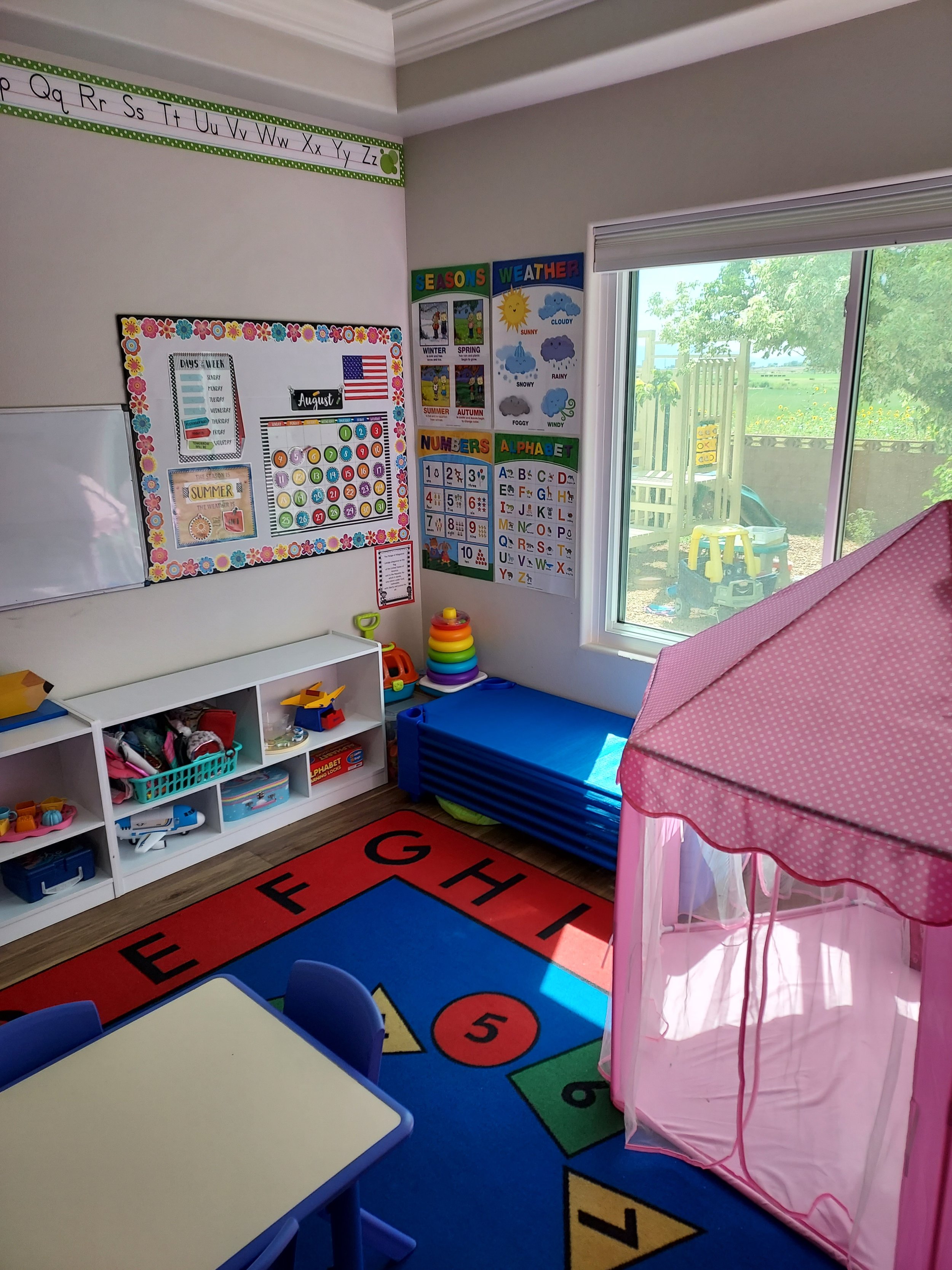 Colorful preschool classroom with educational posters on the wall, a white shelf with toys, a blue foam mat, a red jumper rug, and a pink fabric tent by a window with outdoor toys outside.