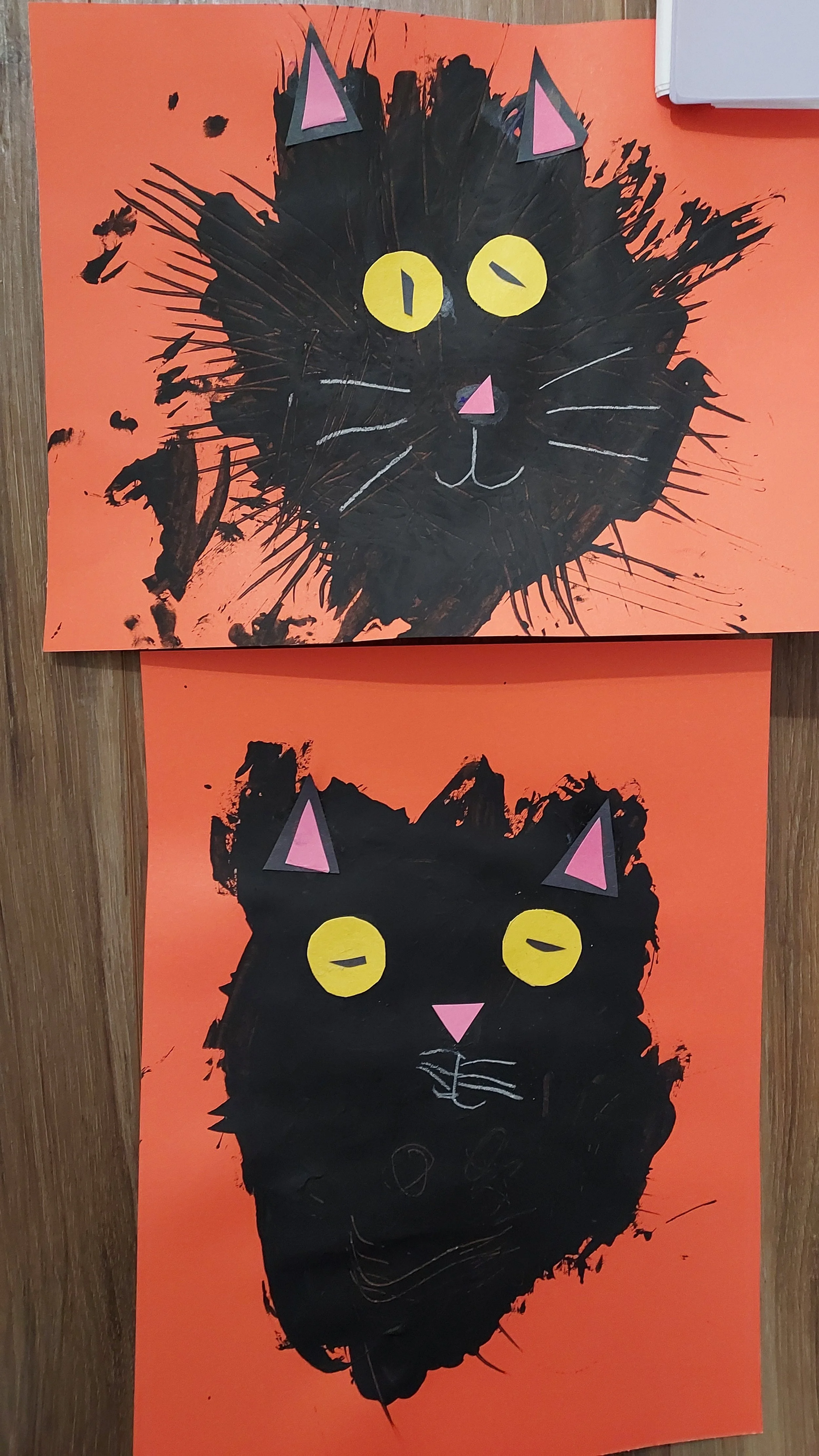 Two children's art projects of black cat faces on orange backgrounds, made with cut-out paper for eyes, nose, and ears, and white lines for whiskers and facial features.