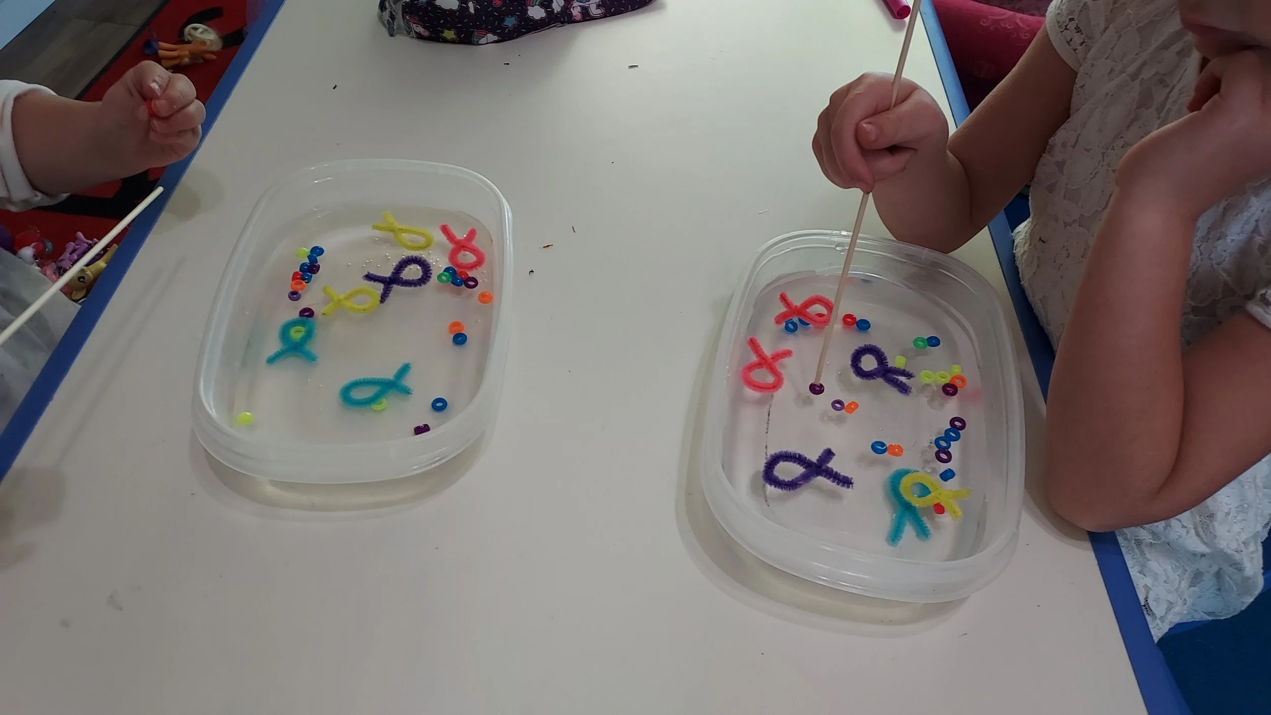 Two children sitting at a table with plastic containers filled with water and colorful pipe cleaners shaped into ribbons and letters, engaging in a sensory activity.