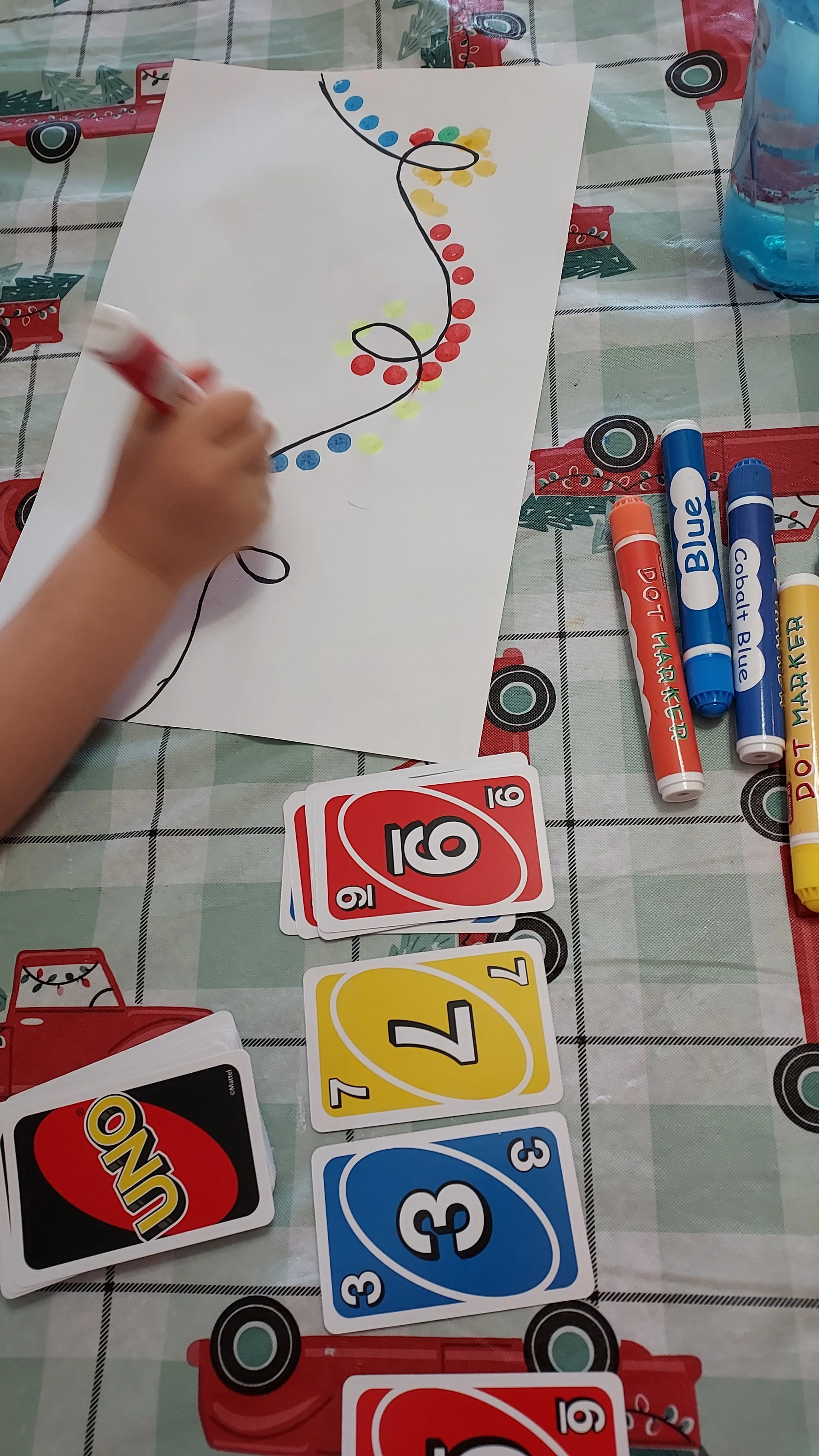 A child is playing Uno card game on a table covered with a holiday-themed tablecloth. The child is drawing or coloring a picture with markers, and there are Uno cards and markers on the table.