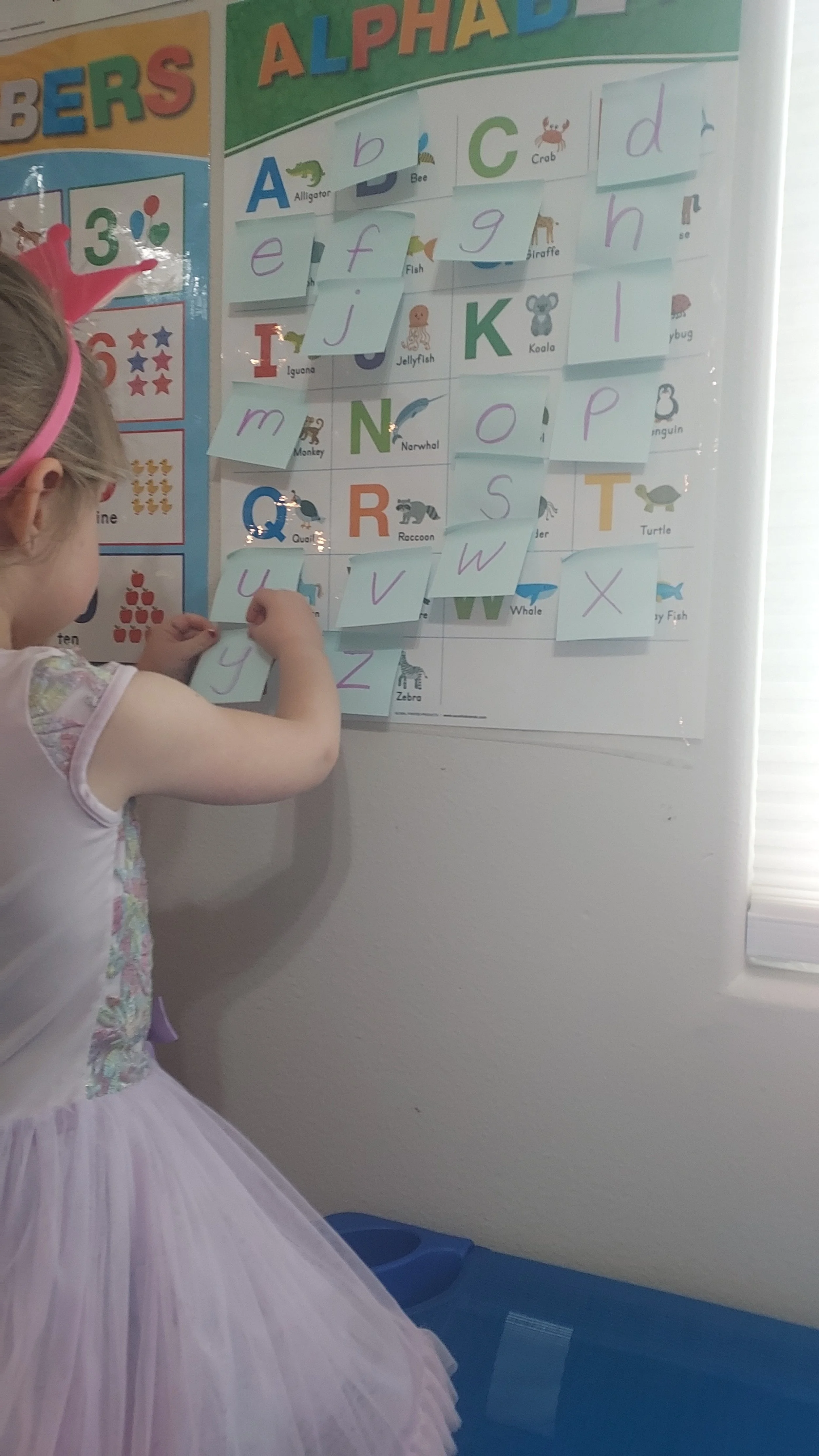 A young girl in a pink princess dress placing letter cards on a classroom alphabet poster, which features each letter with corresponding images and words.