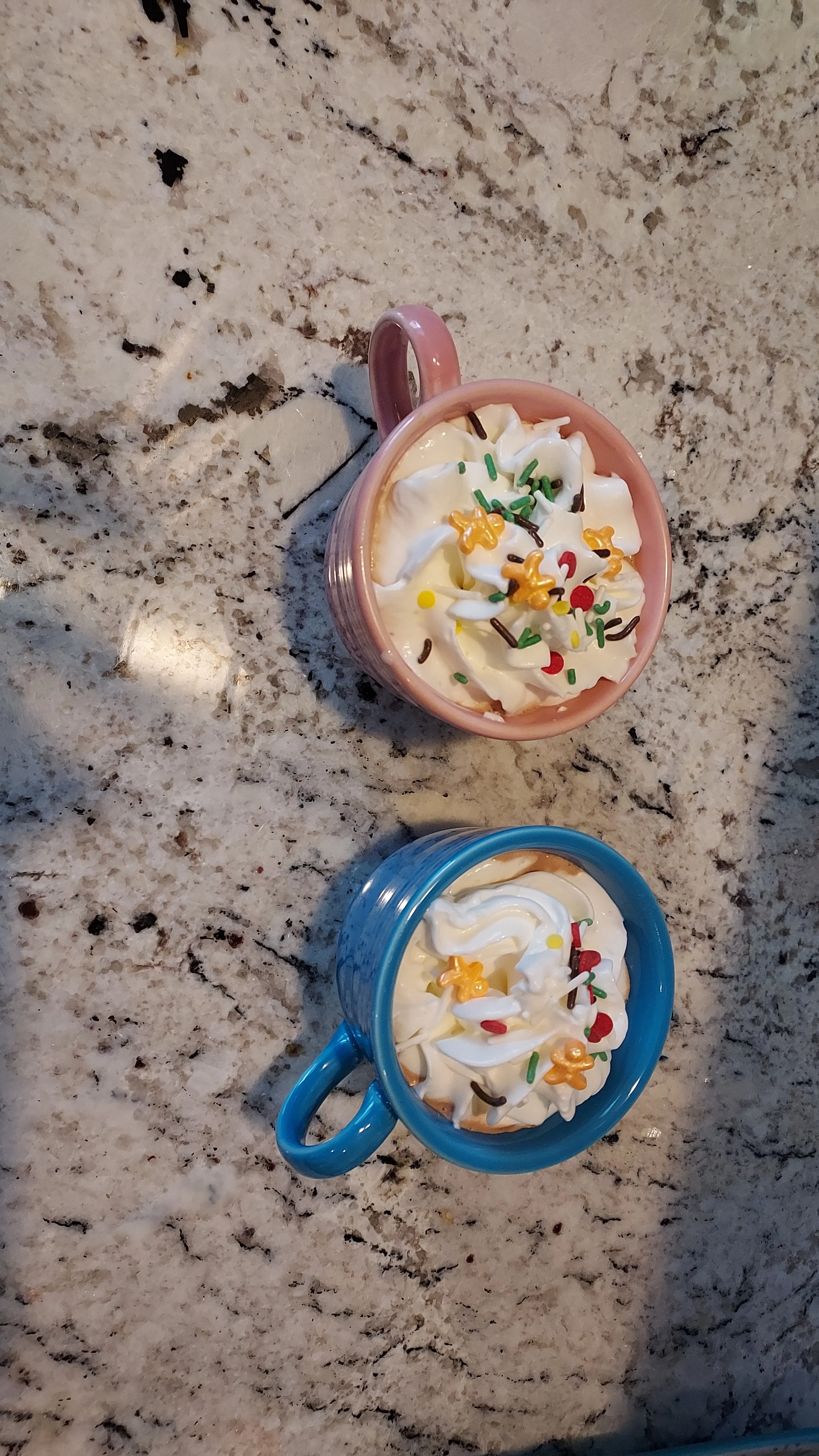 Two mugs of whipped cream topped with colorful sprinkles on a granite countertop.