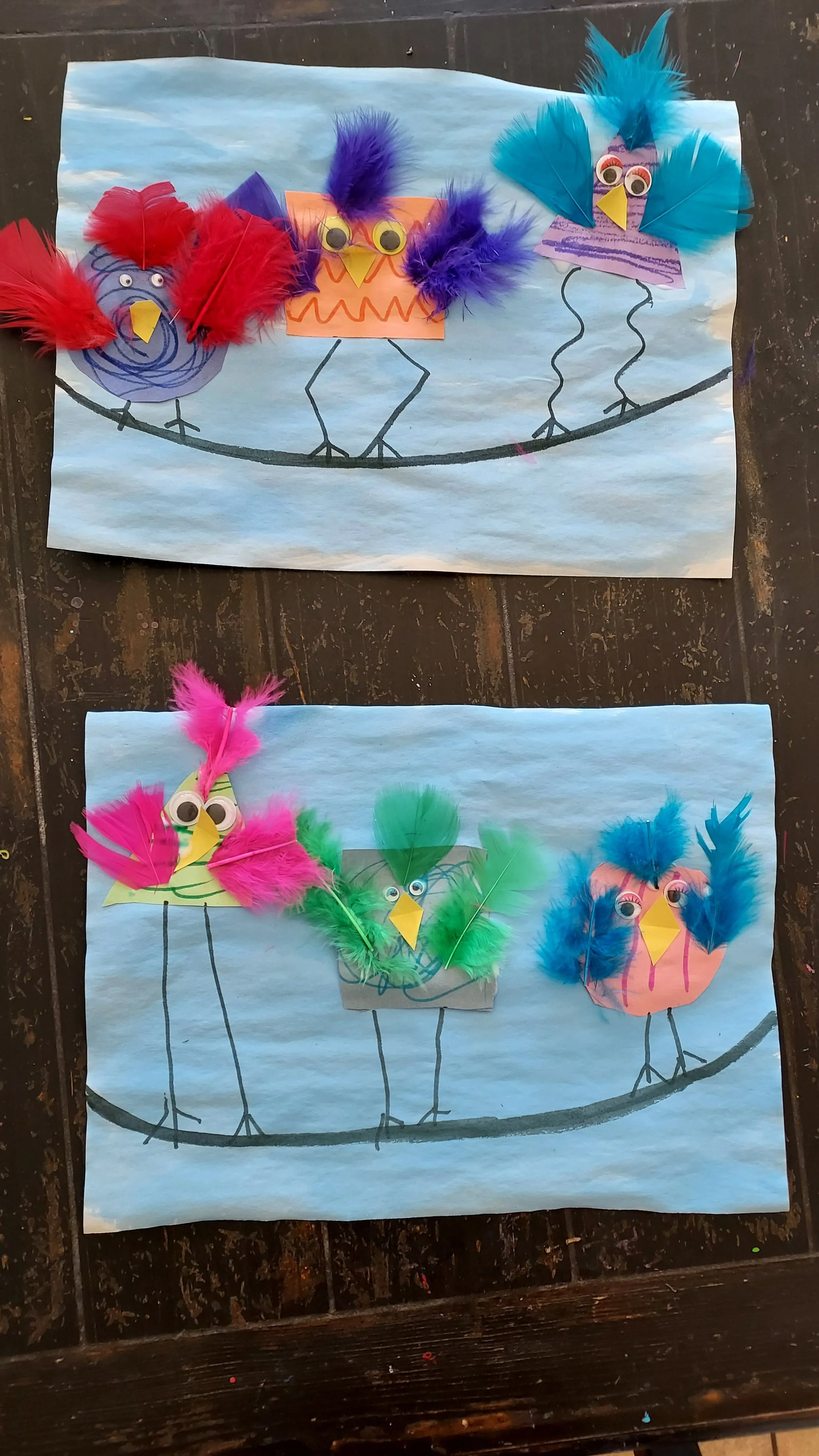 Colorful paper crafts of birds with feather plumes, googly eyes, and drawn bodies, arranged on blue construction paper with a black line sketch of a branch.