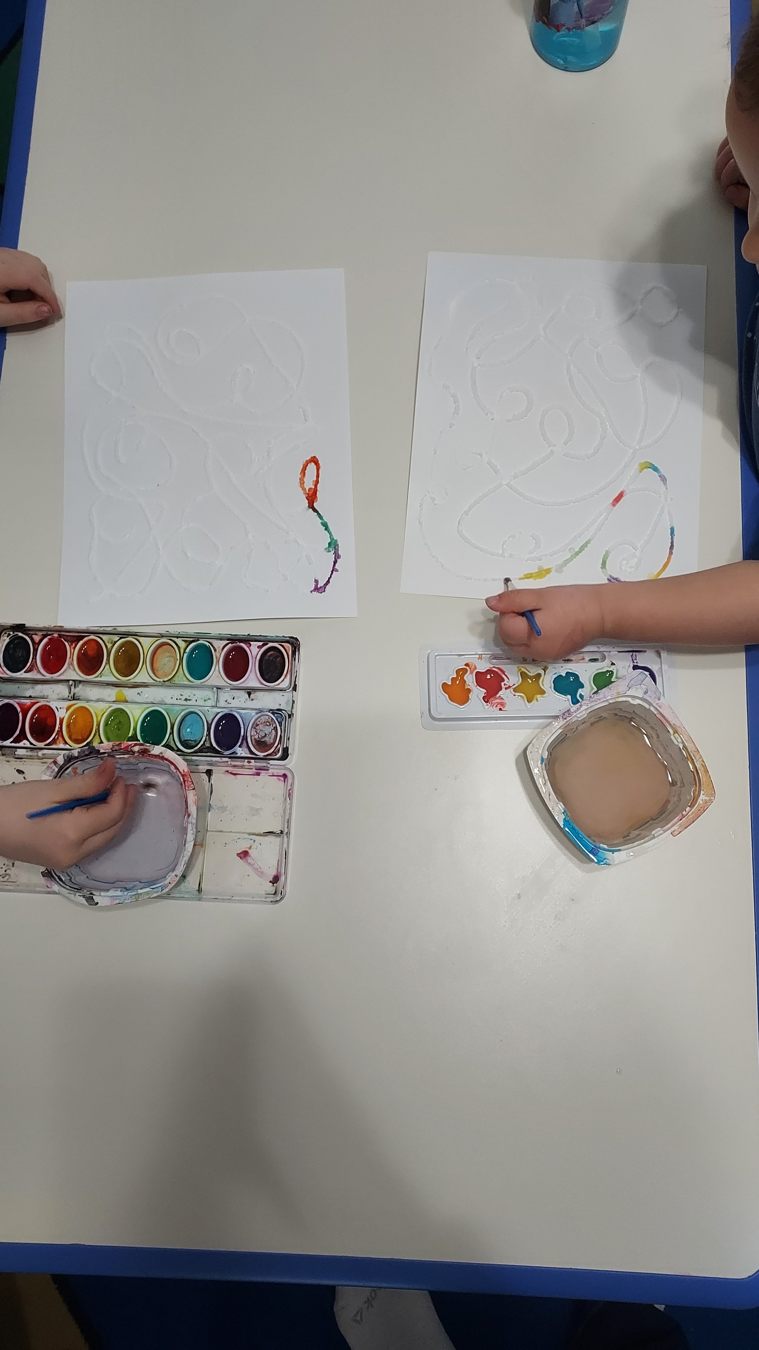 Children painting with watercolors on white paper; one child's hand is visible, applying colors, and another child's face is partially visible at the right.