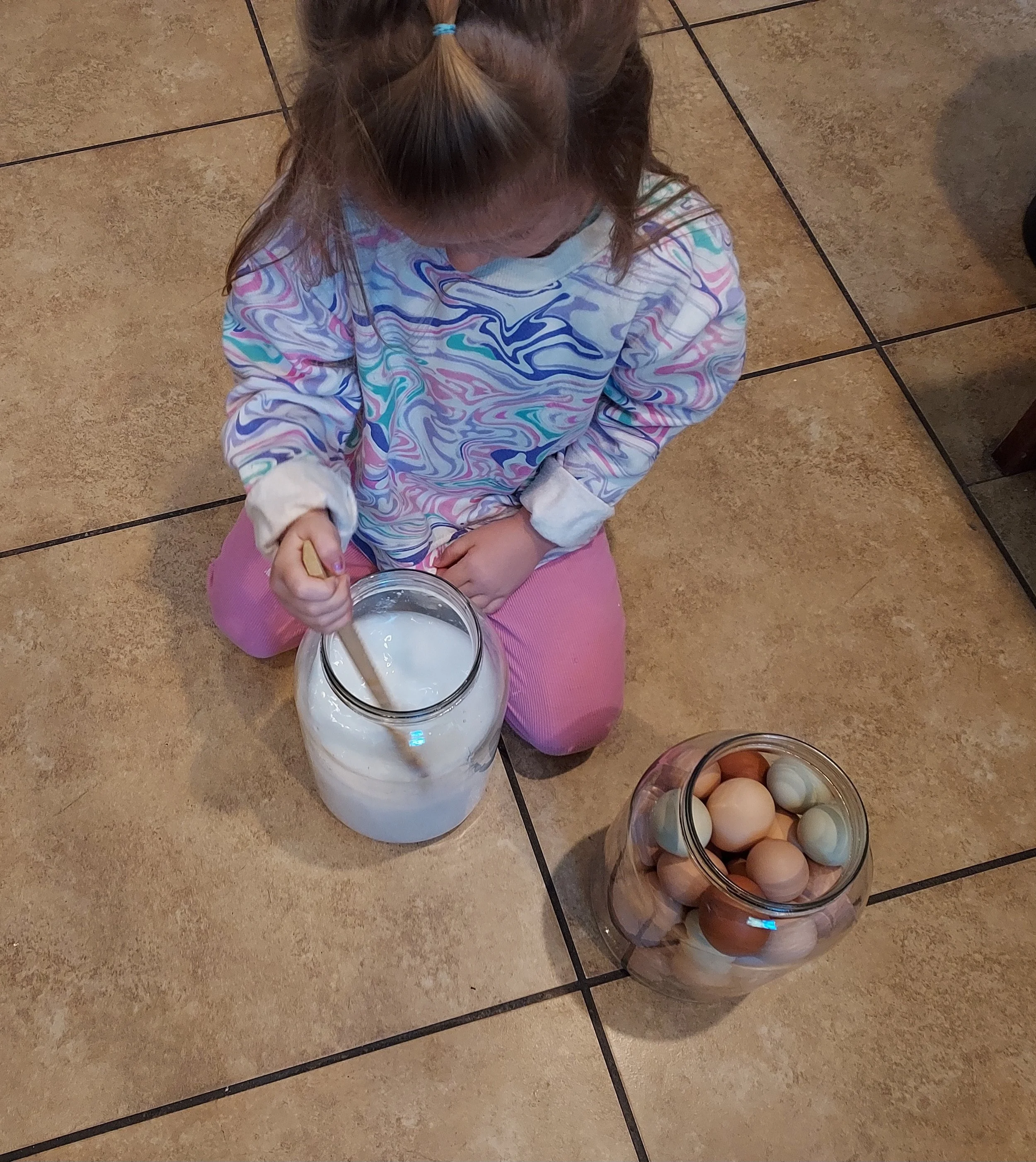 A young girl sitting on a tiled floor, stirring a large glass jar of milk with a wooden spoon. Next to her is a jar filled with various colored eggs.