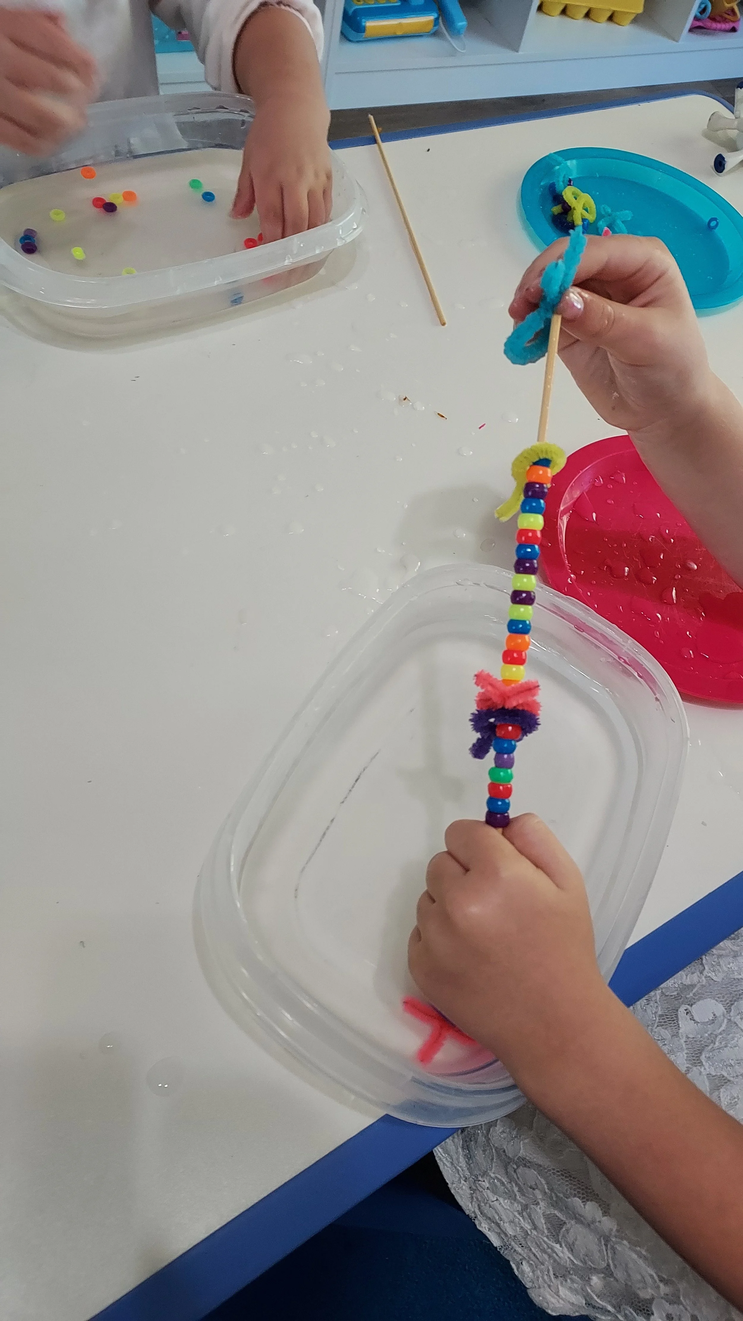 Child's hands threading colorful beads and pipe cleaners to make a craft in a white table, with water, bowls, and craft supplies around.