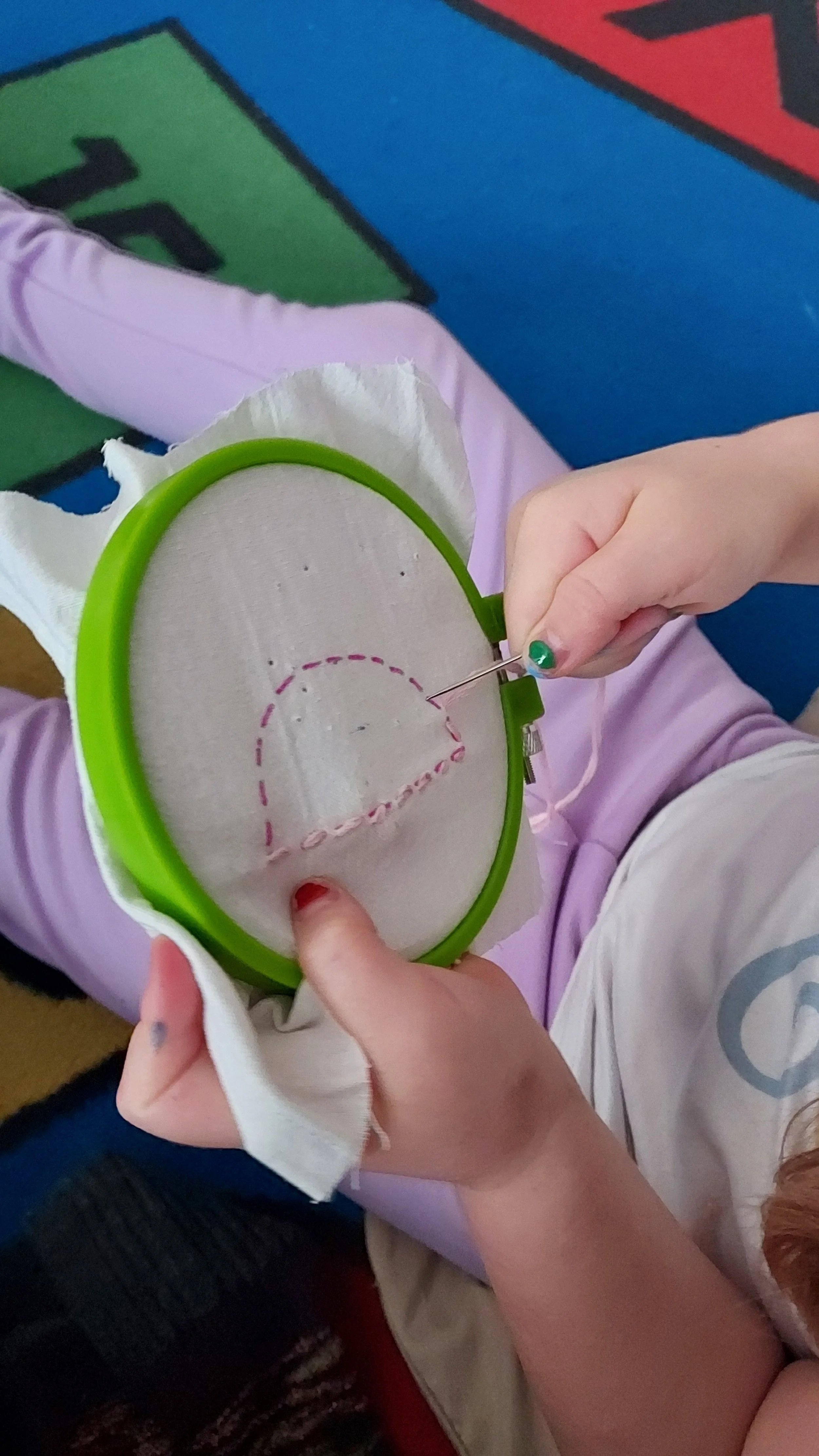 A person is embroidering on a piece of fabric held in a green embroidery hoop, with the focus on their hands and the embroidery needle. The fabric has a pink dashed outline of a heart shape being stitched.