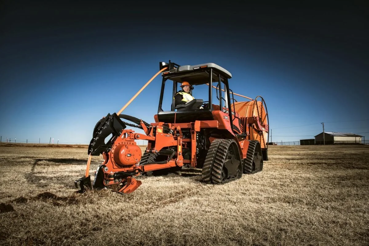 A person in safety gear operating a large orange tracked machine on a dry grassy field, with a clear blue sky and distant buildings in the background.