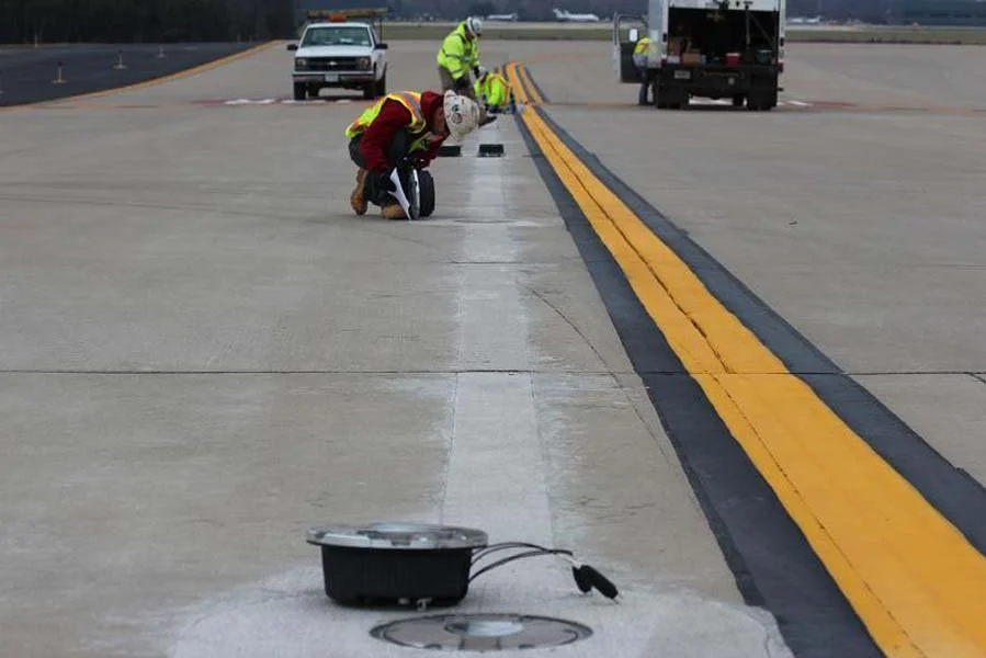Workers repainting yellow and black safety lines on an airport runway or tarmac in an open outdoor setting during daytime.