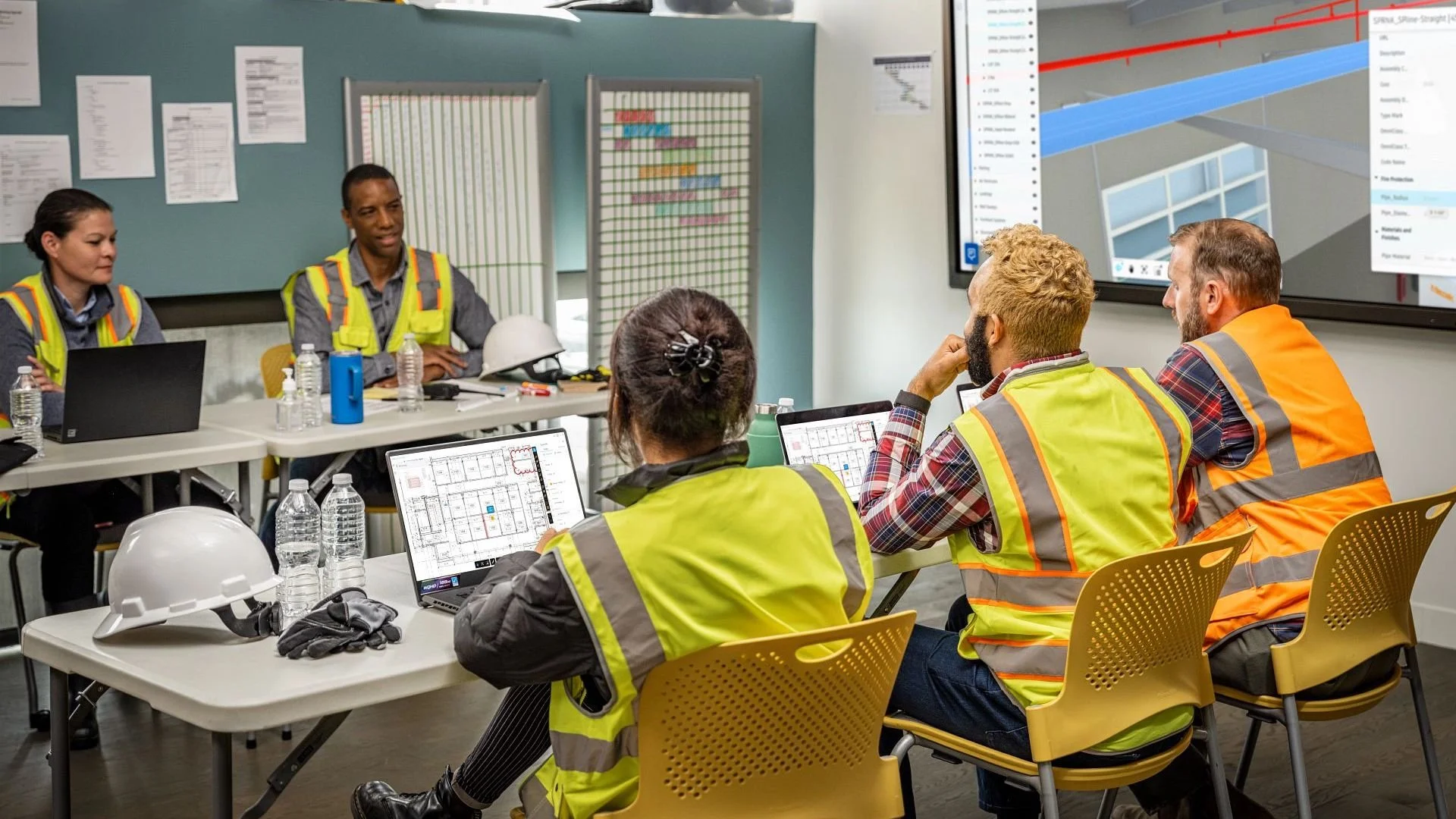 A group of construction professionals in safety vests attending a meeting in a conference room with construction plans on laptops and large screens showing charts and graphs.