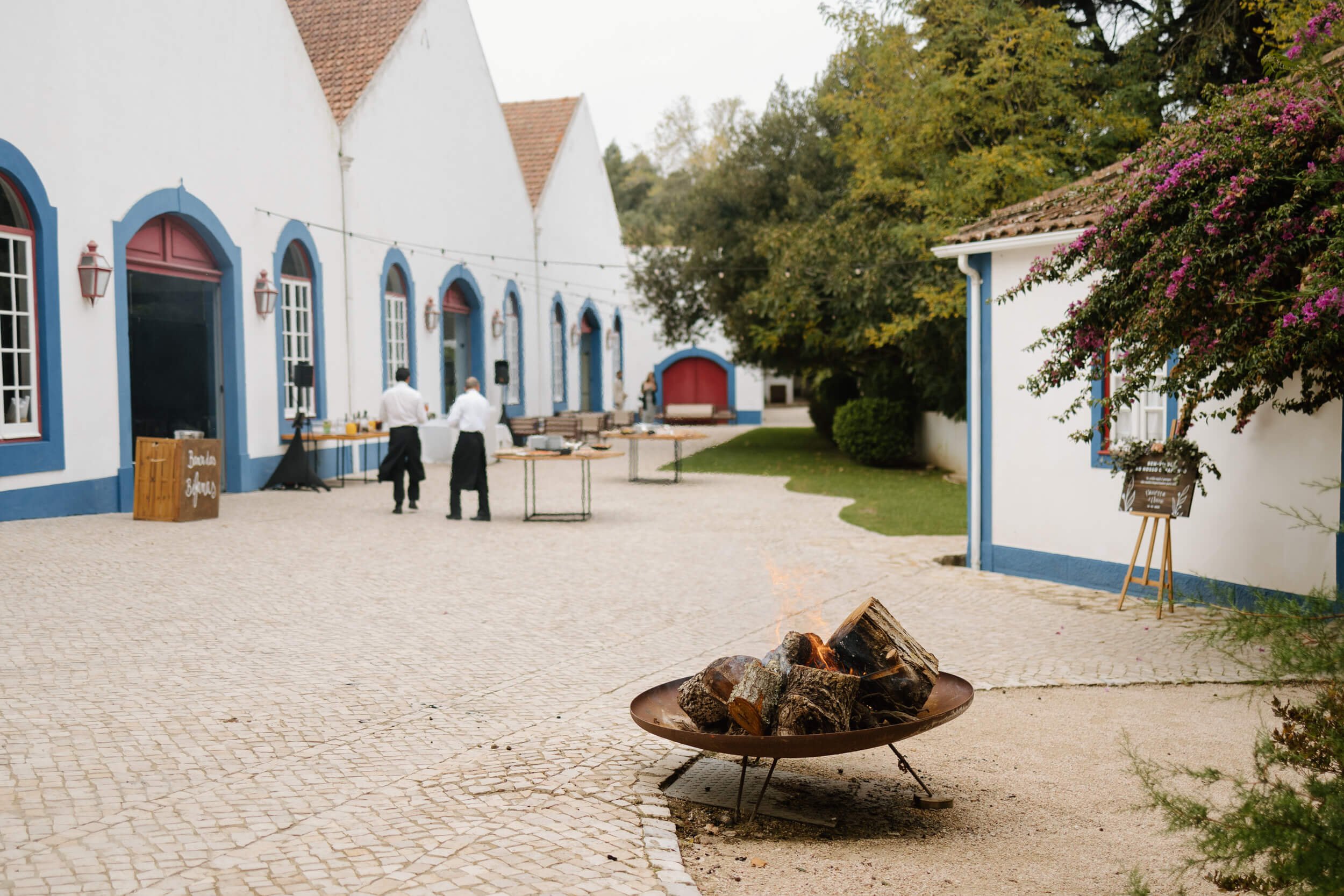 A courtyard with cobblestone pavement, two people in white shirts and black aprons setting up a buffet, a fire pit with burning logs in the foreground, and white buildings with blue trim and red-tiled roofs, surrounded by trees and flowering bushes.