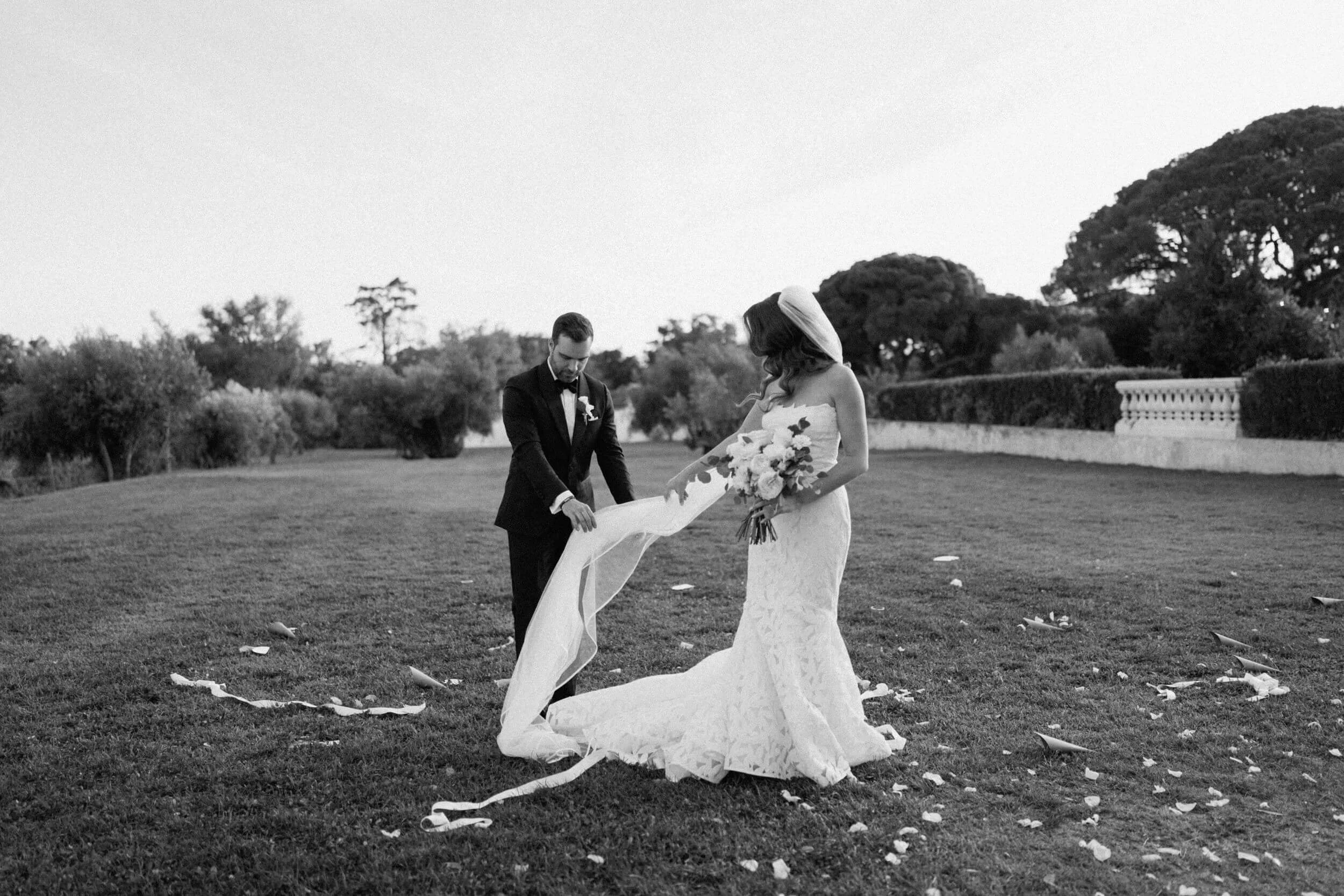 A black and white photo of a bride and groom on a grassy field, with the groom holding the bride's train. Flower petals are scattered on the ground, and trees and a white balustrade are in the background.