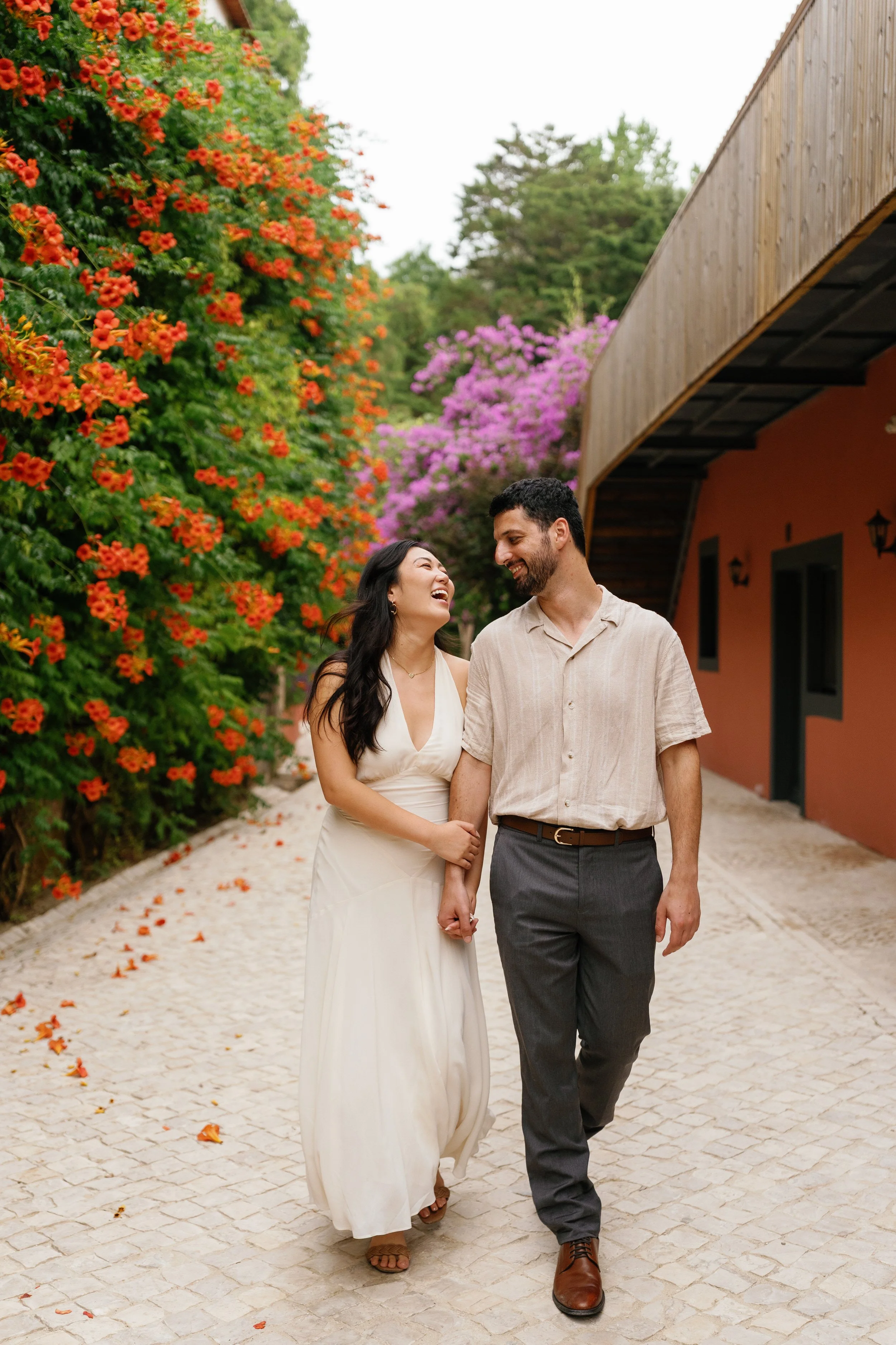 A smiling couple holding hands and walking on a cobblestone path surrounded by colorful flowers and trees, with a building on the side.