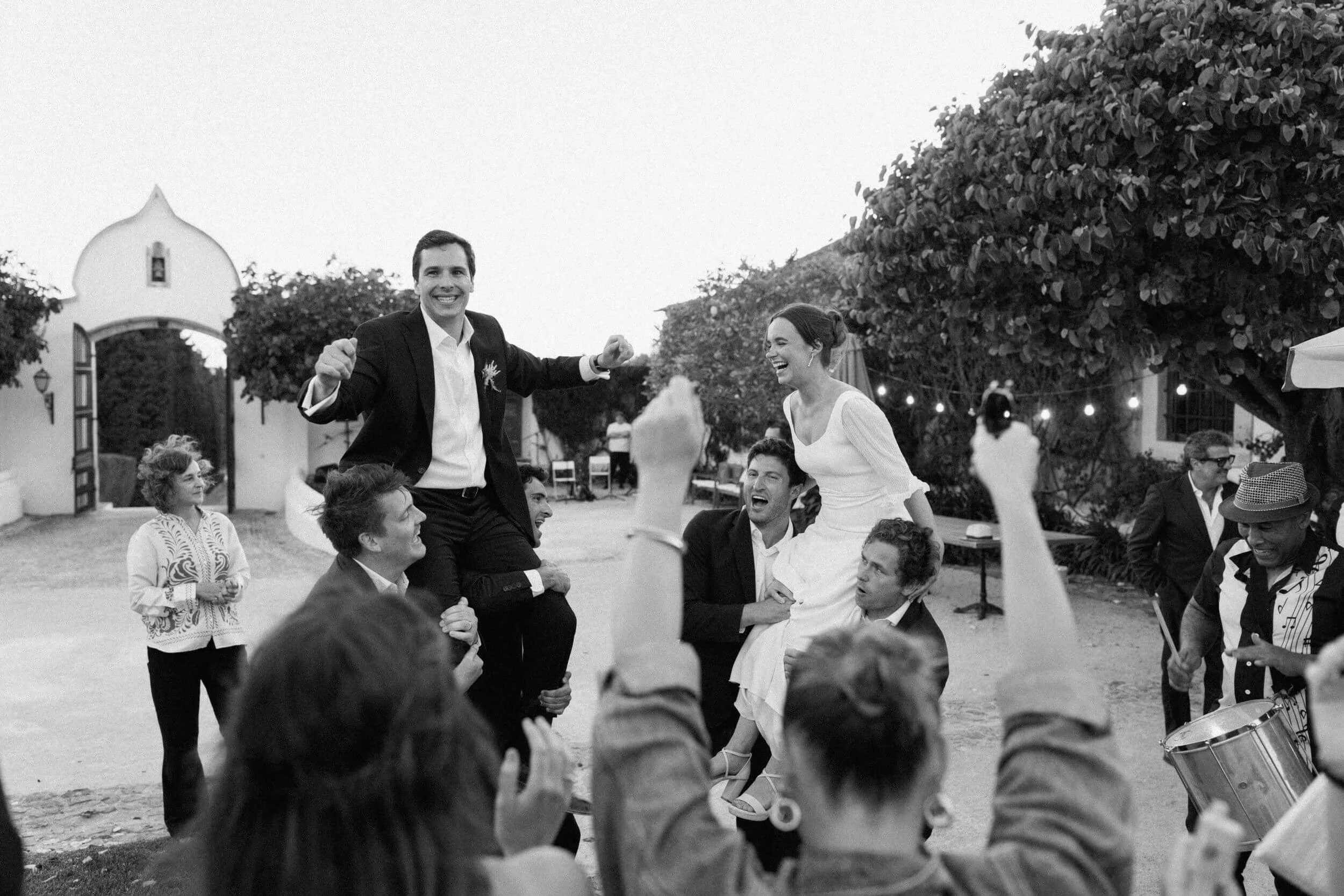 People celebrating at a wedding reception outdoors, with one person lifted on others' shoulders and a woman laughing, during evening under string lights.
