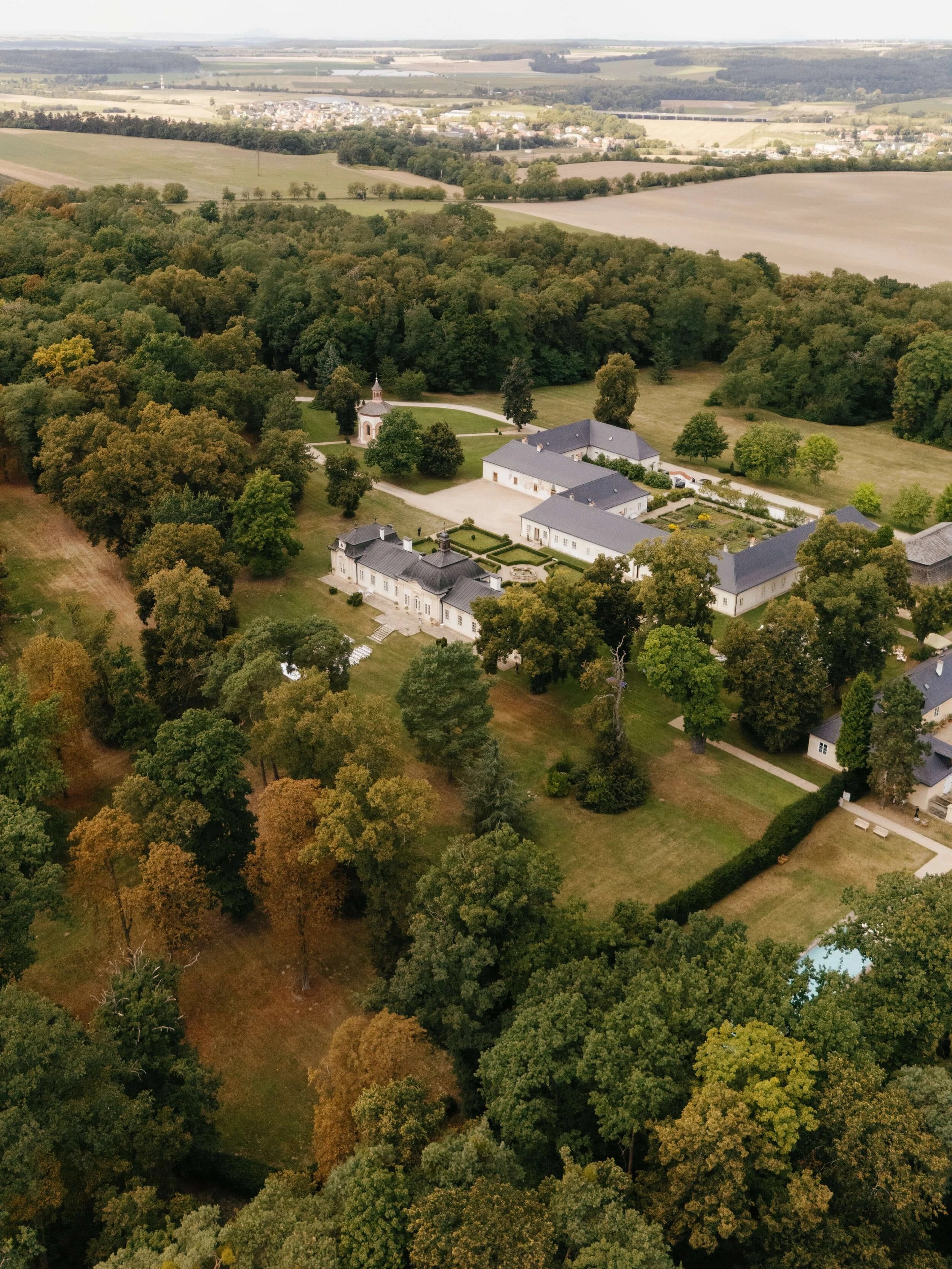 Aerial view of a large estate with white buildings, a well-maintained garden, and surrounding trees in a rural landscape.