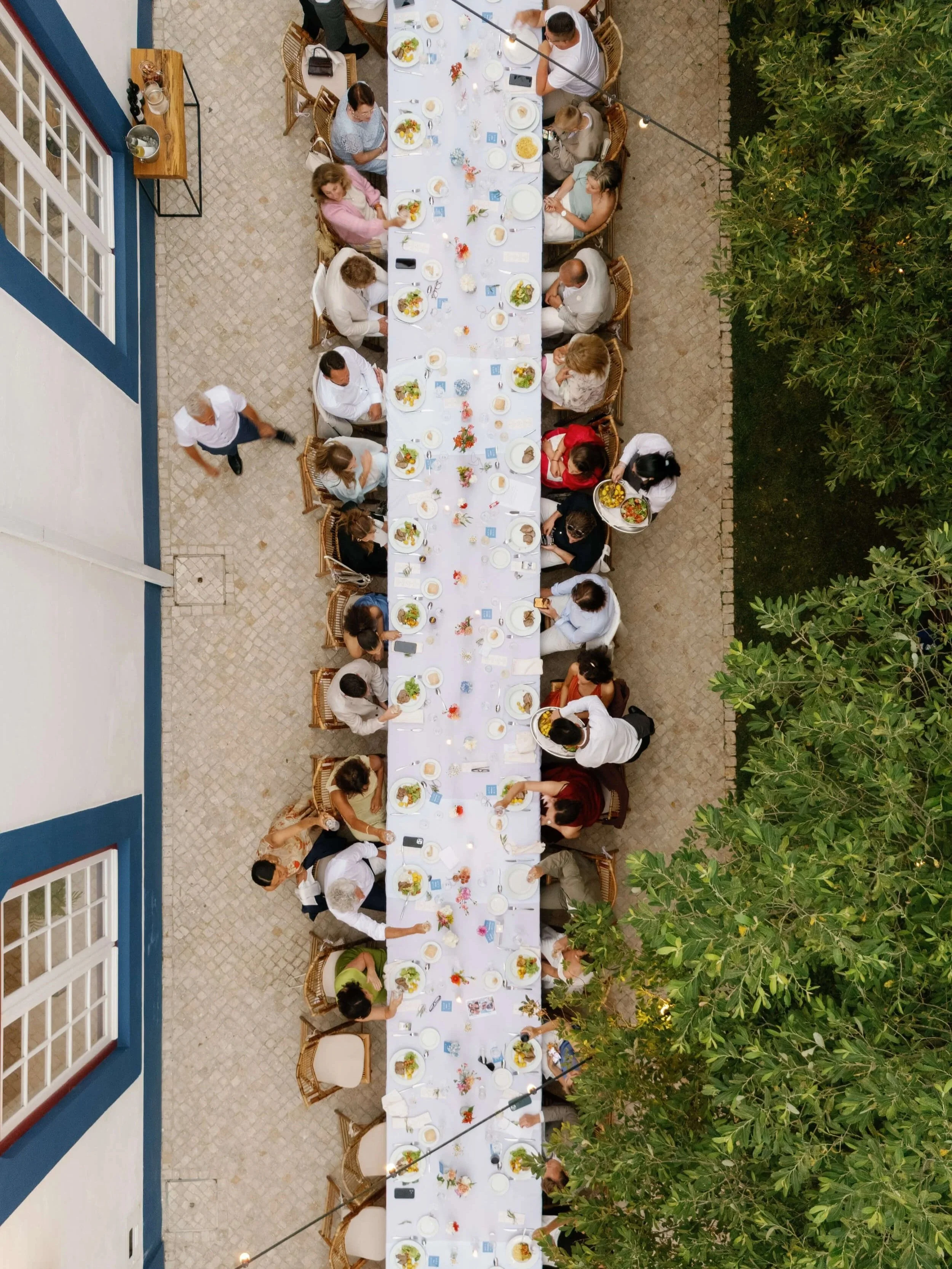 Overhead view of a long outdoor dining table with people eating, with a building wall on the left and greenery on the right. The table is decorated with flowers and has plates of food and drinks.