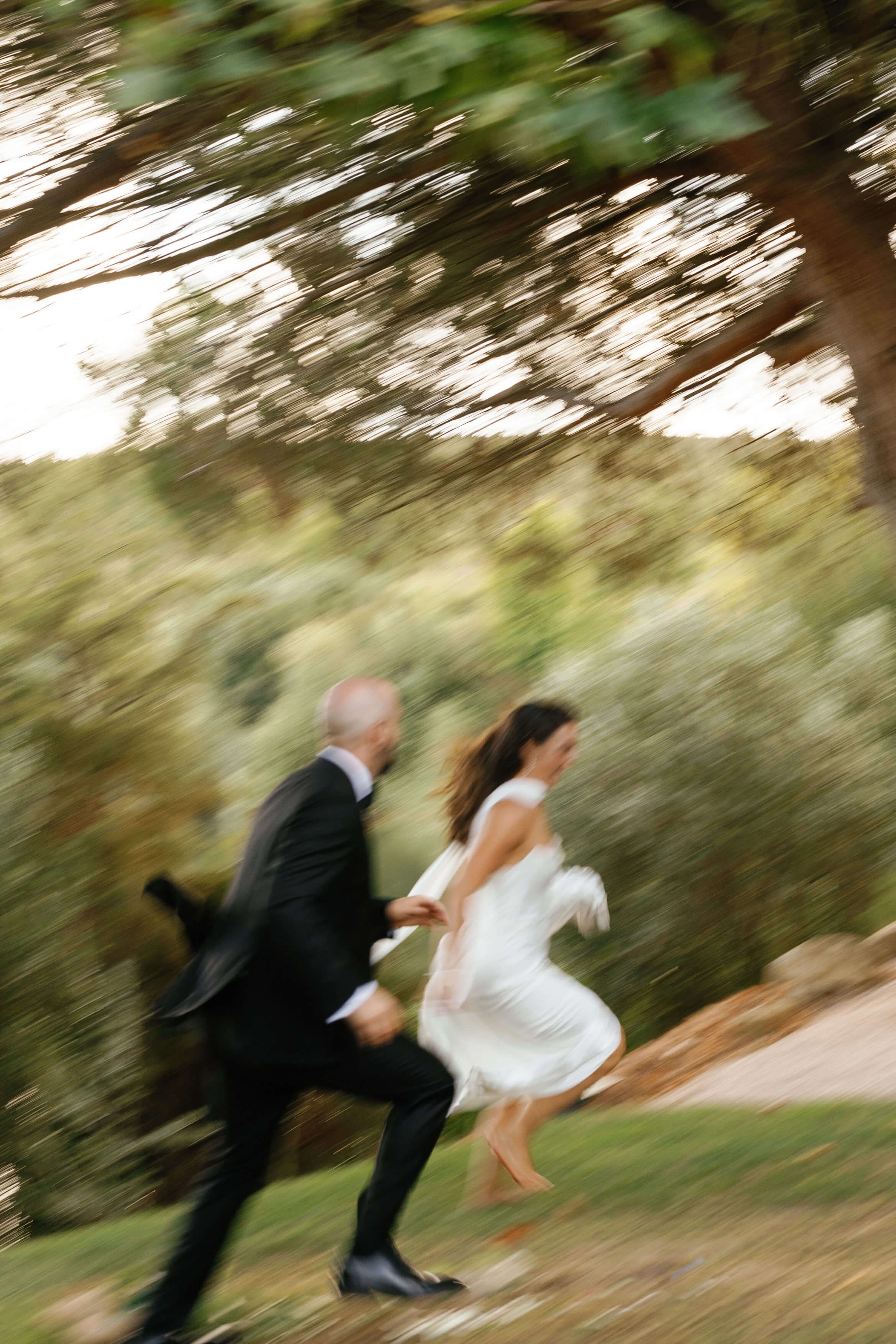 A couple dressed in wedding attire running outdoors on a grassy area with trees, with a motion-blurred background.