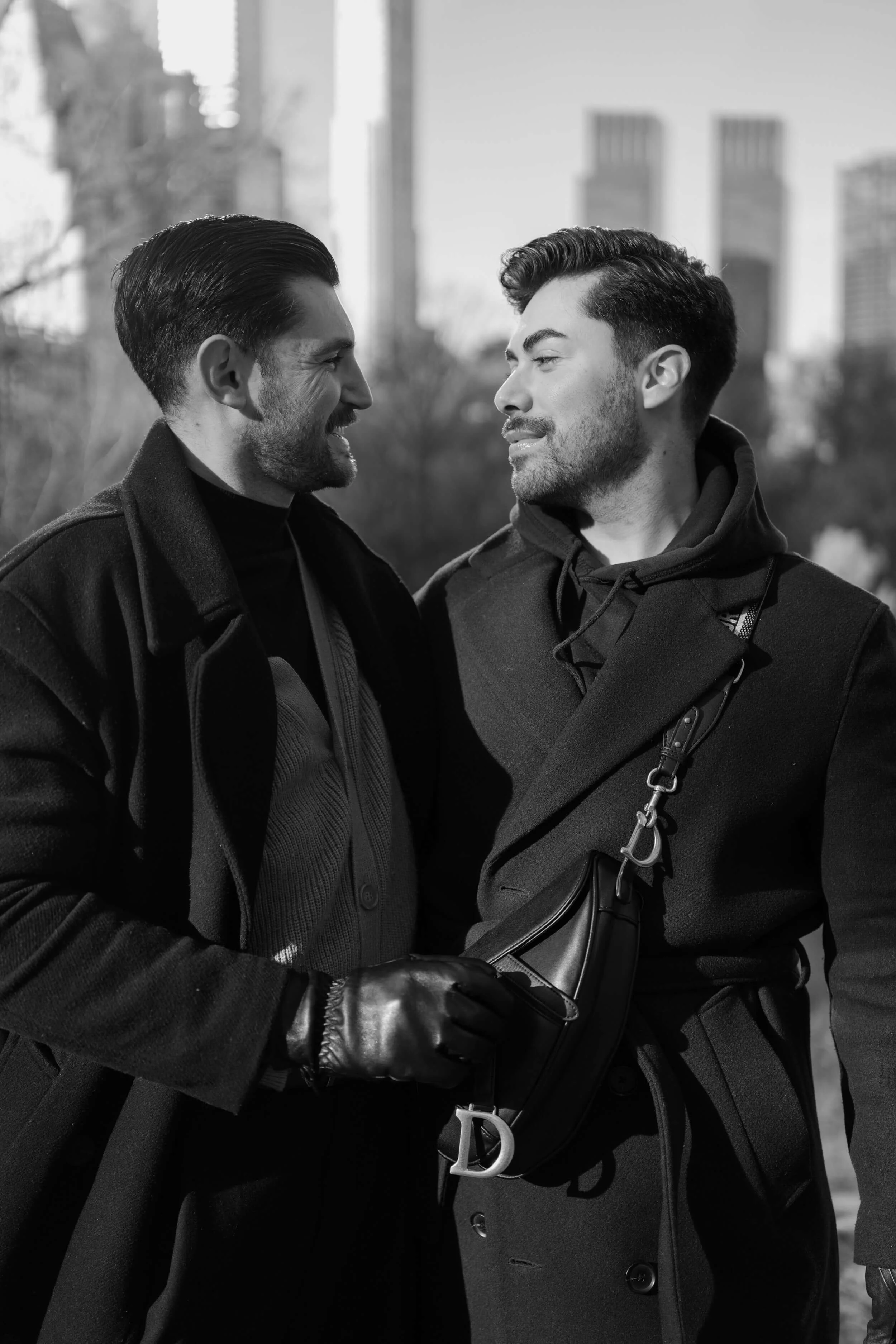 Two men are smiling and looking at each other outdoors in a city park, dressed warmly in coats and gloves, with buildings in the background.