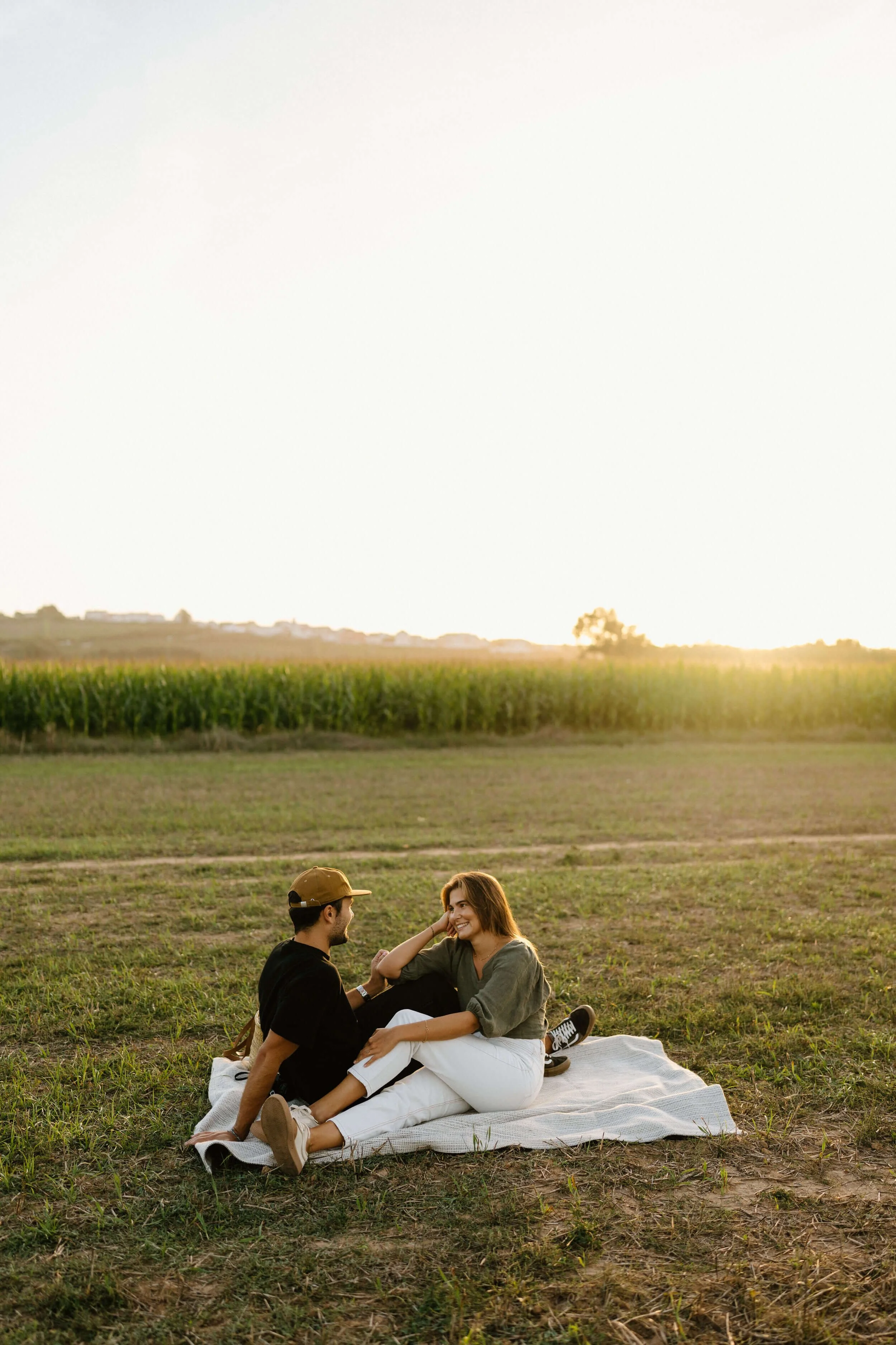 A young couple sitting on a blanket in an open field during sunset, smiling and enjoying each other's company.