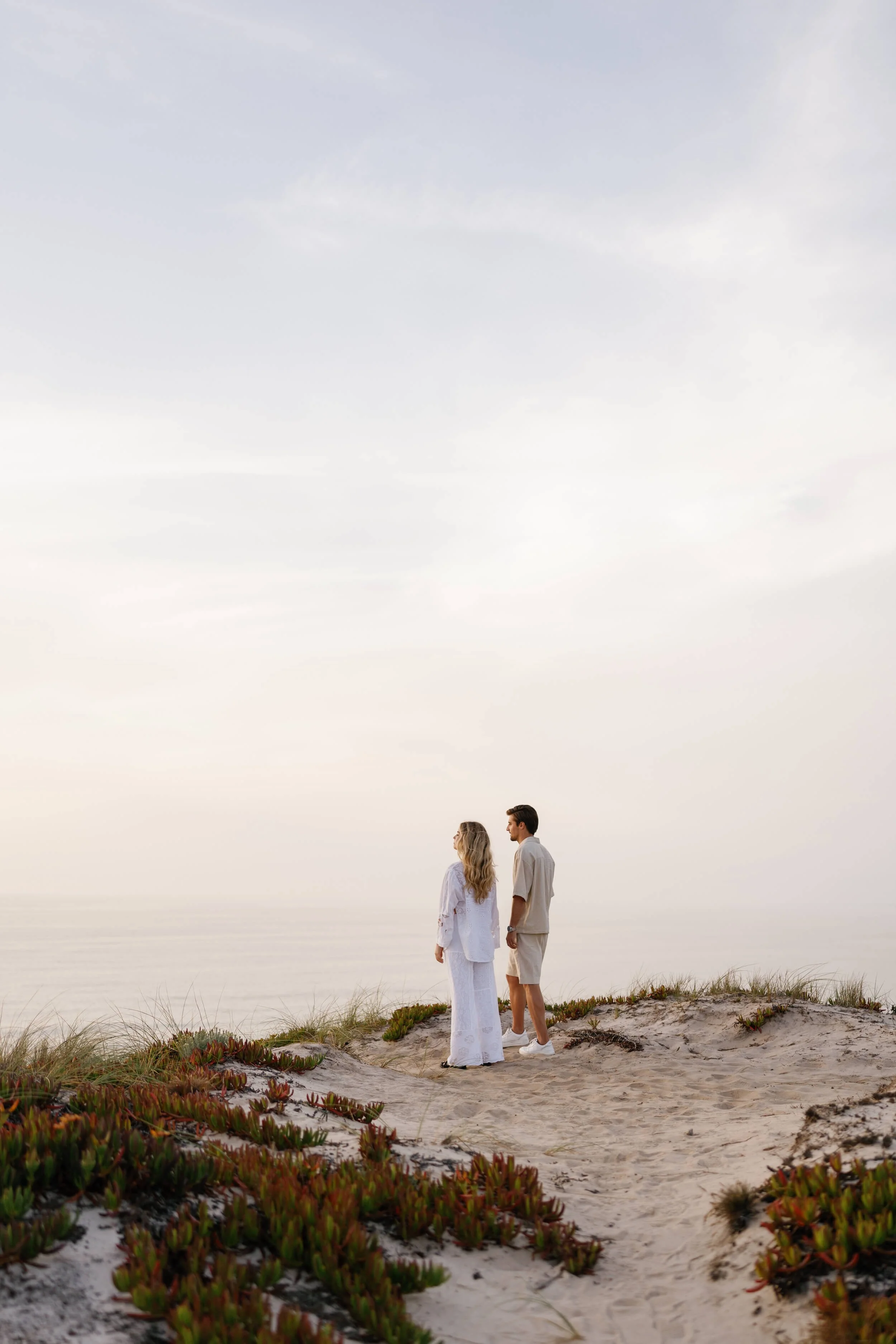 A couple standing on a sandy beach with some vegetation, facing each other, under a cloudy sky.
