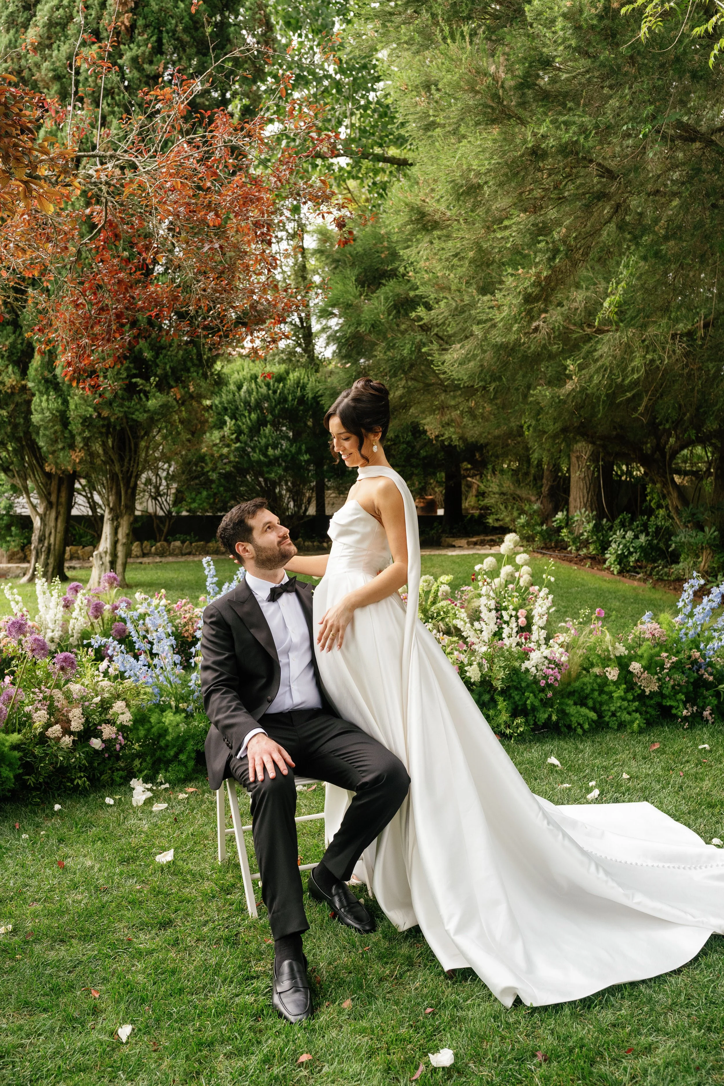 A wedding couple in a lush garden with colorful flowers and green trees. The bride is in a white wedding dress, standing and smiling down at the groom, who is seated in a black tuxedo and looking up at her.