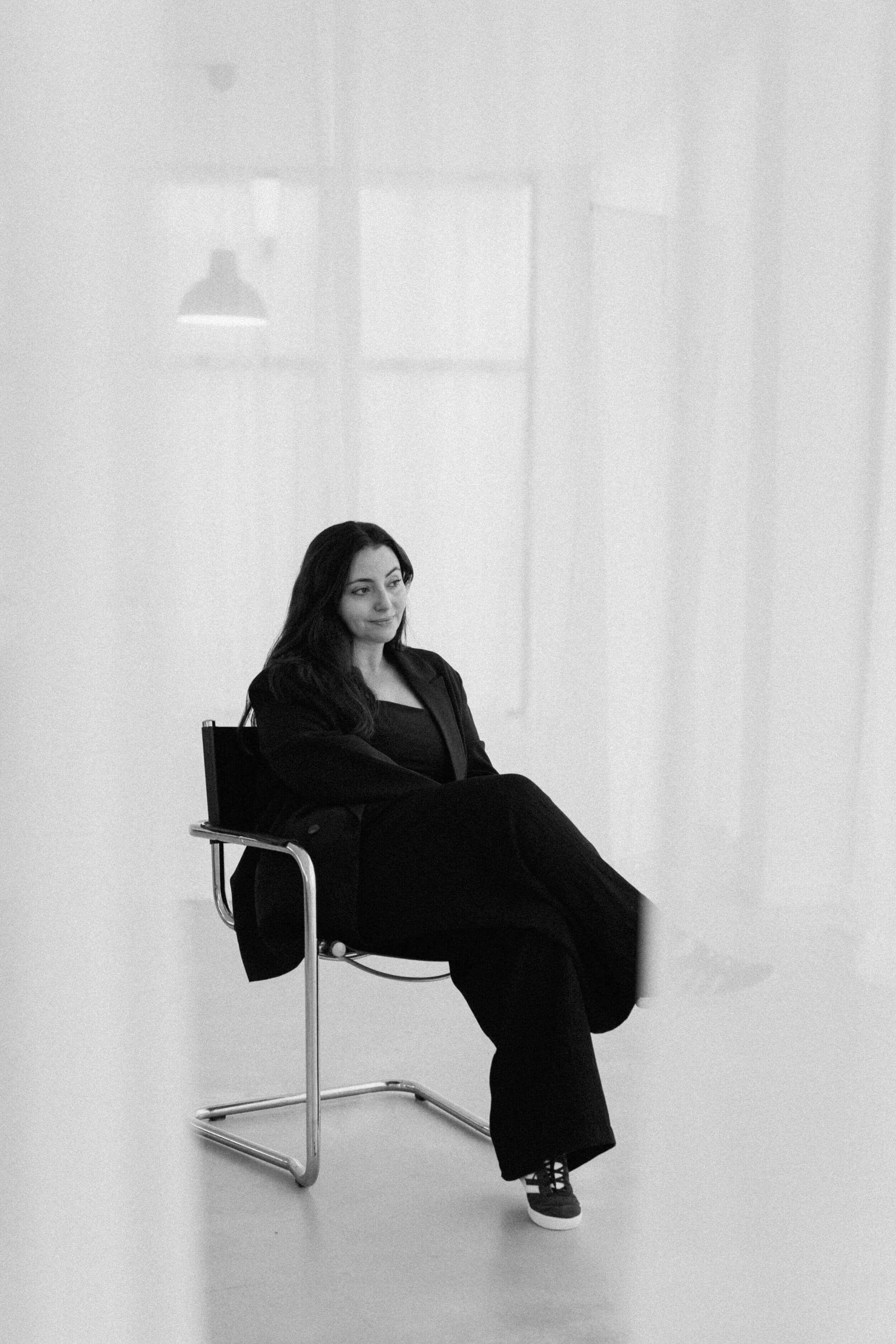 A woman with long dark hair smiling, looking to her side, while seated down in a chair in the middle of a photography studio with white curtains all around