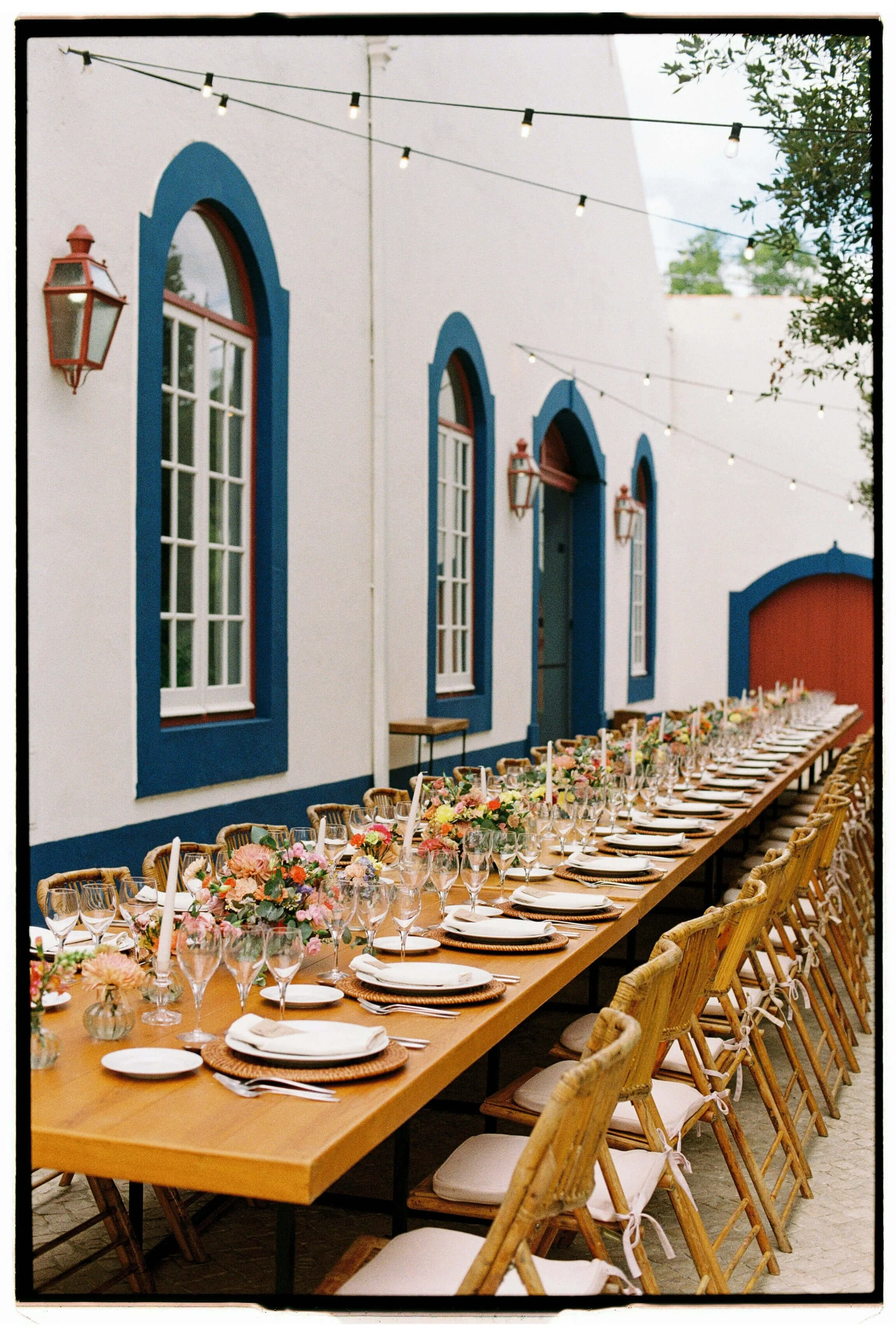 Long dining table set for an event with floral centerpieces, candles, and place settings, outdoors in front of a white building with blue-trimmed windows and string lights overhead.