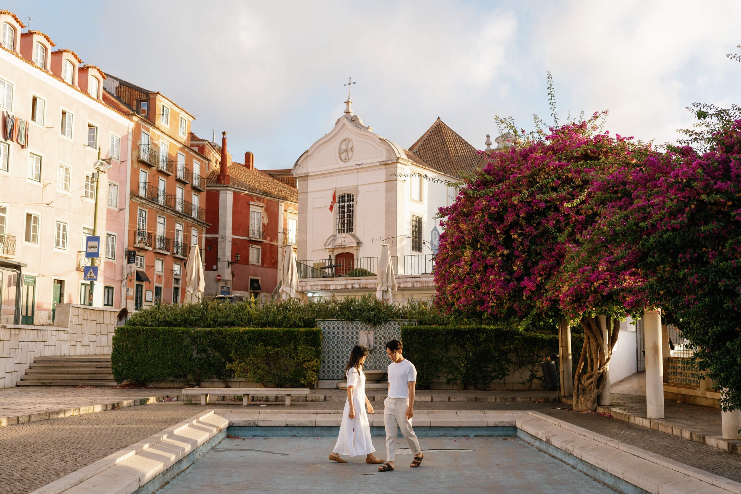 A couple dancing in an outdoor plaza with a large pink flowering tree, a church, and colorful buildings in the background during the daytime.