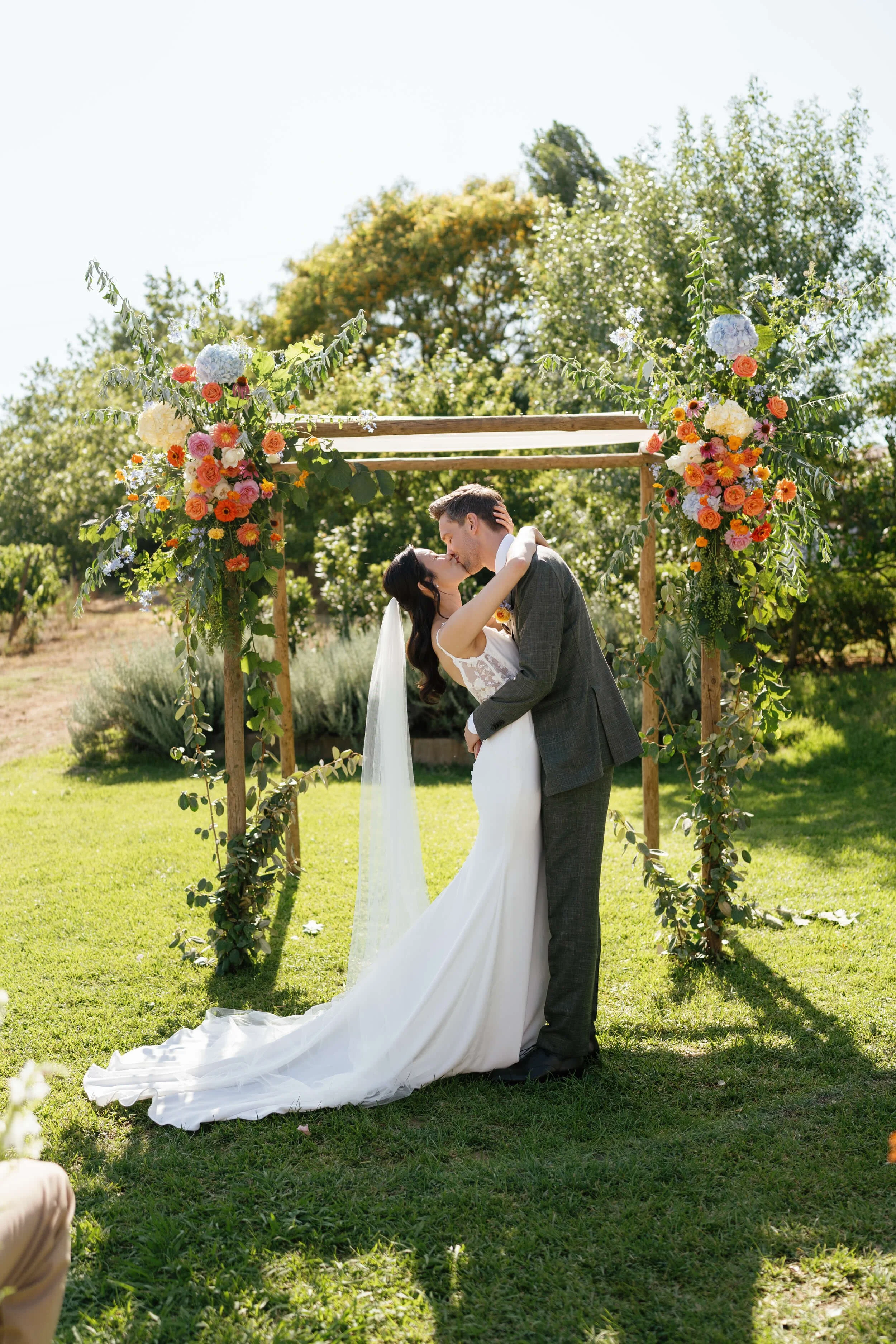 A bride and groom kissing under a wooden floral arch on their wedding day outdoors in Quinta de Sant'Ana do Gradil, Mafra