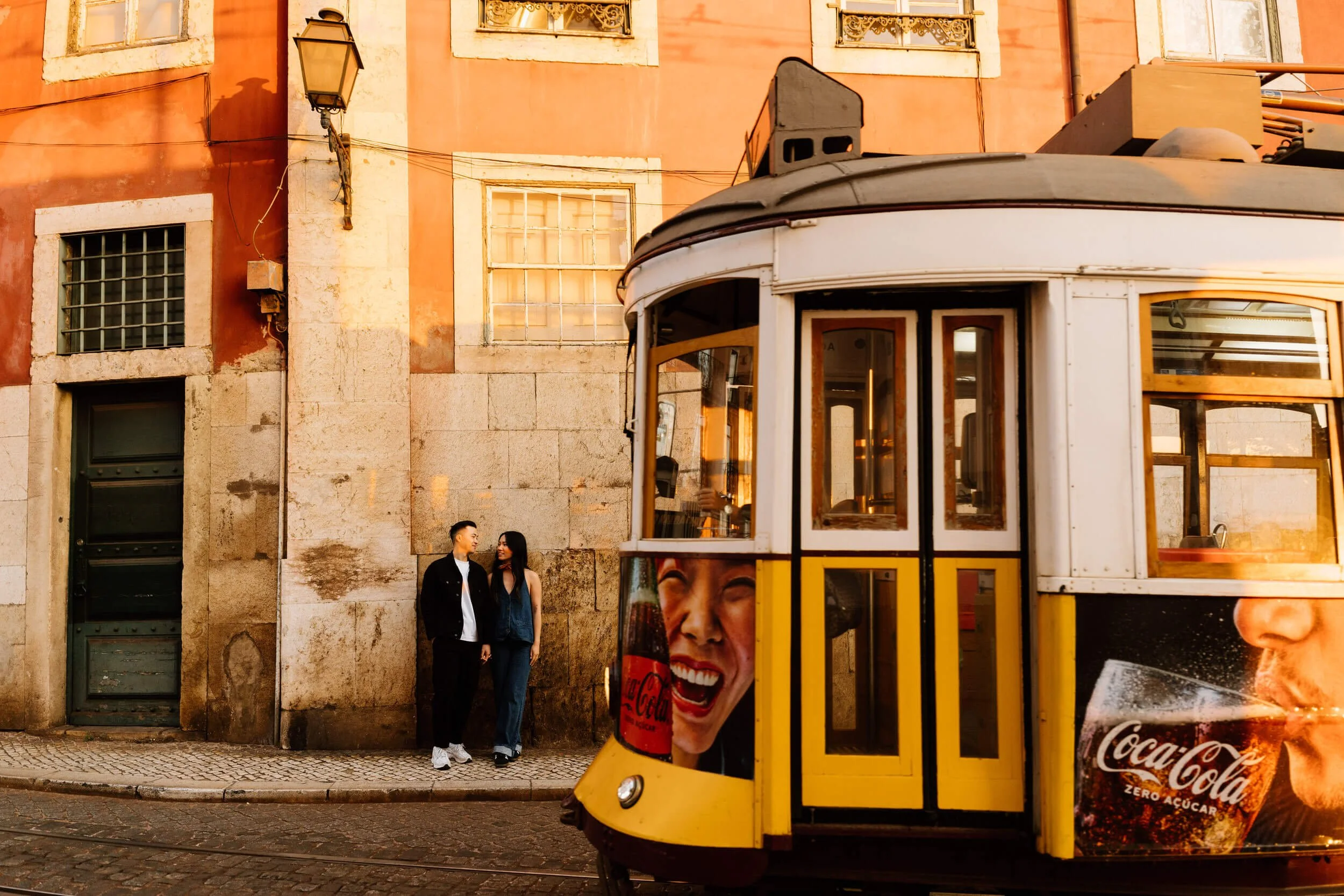 A Lisbon yellow tram, with a smiling woman and a man holding hands standing on the sidewalk in front of an old building at sunset.
