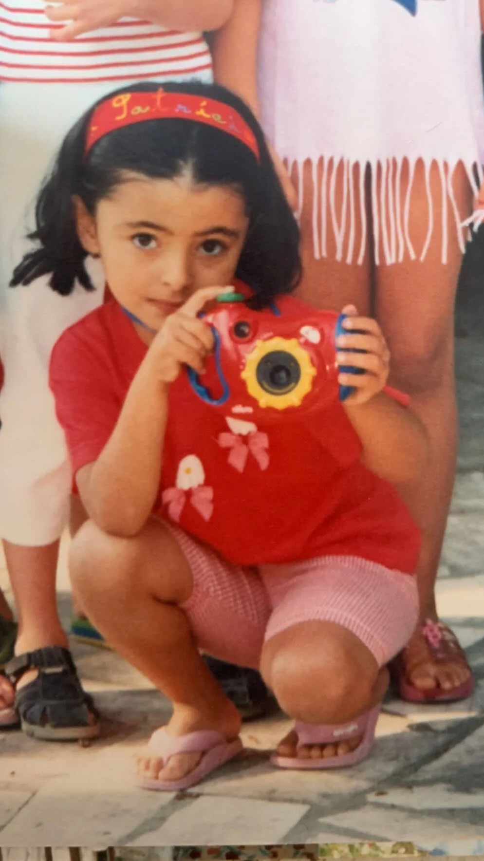 A young girl with black hair and a red headband holding a red toy camera, crouching on a tiled floor, with other children partially visible behind her.
